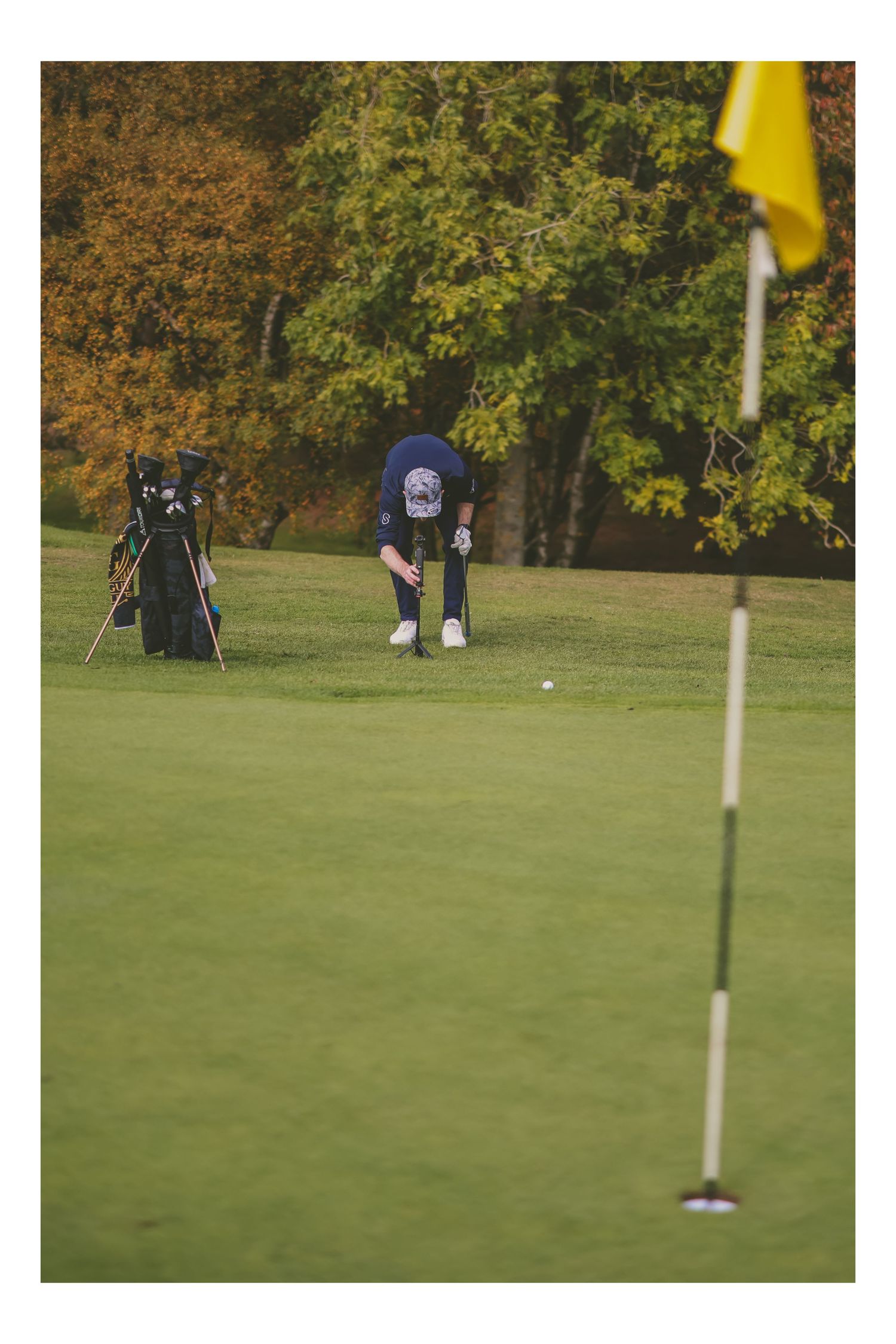 Golfer putting on a green near yellow flag with golf bag in background amid autumn foliage.