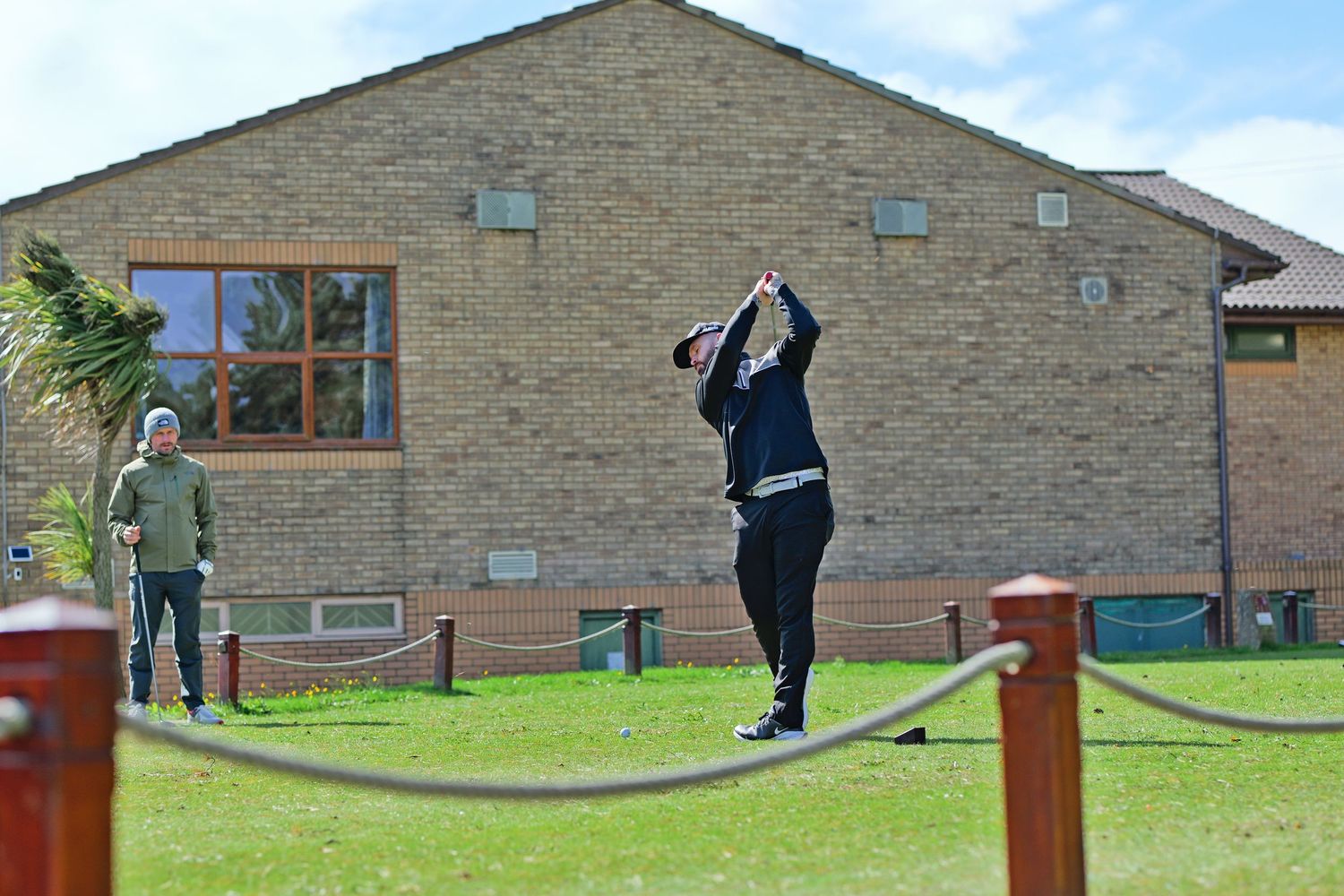 Golfer completing a swing near a brick clubhouse with a spectator watching nearby.