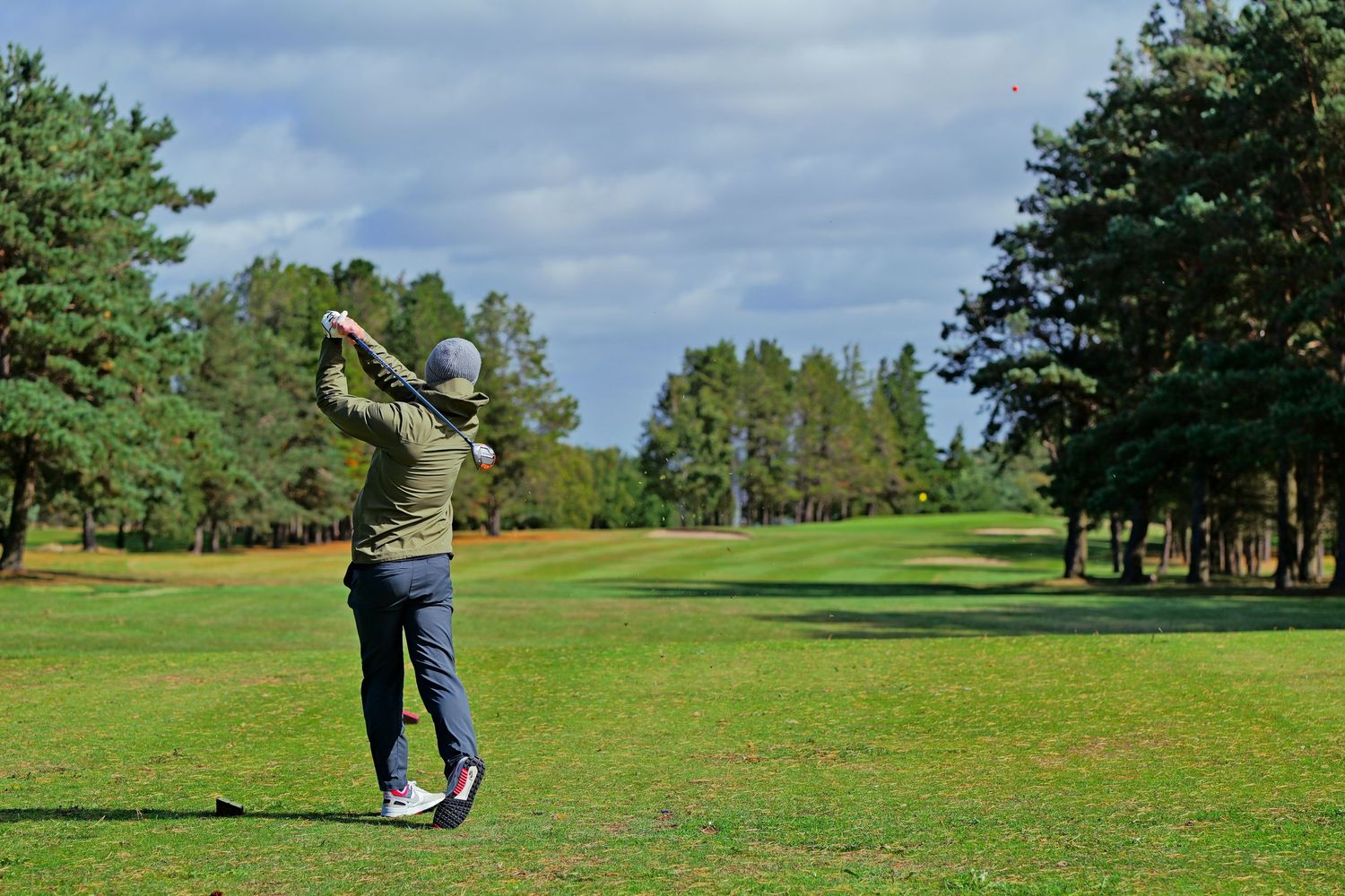 Golfer in olive jacket and jeans finishing a drive on a sunny green golf course lined with pine trees.