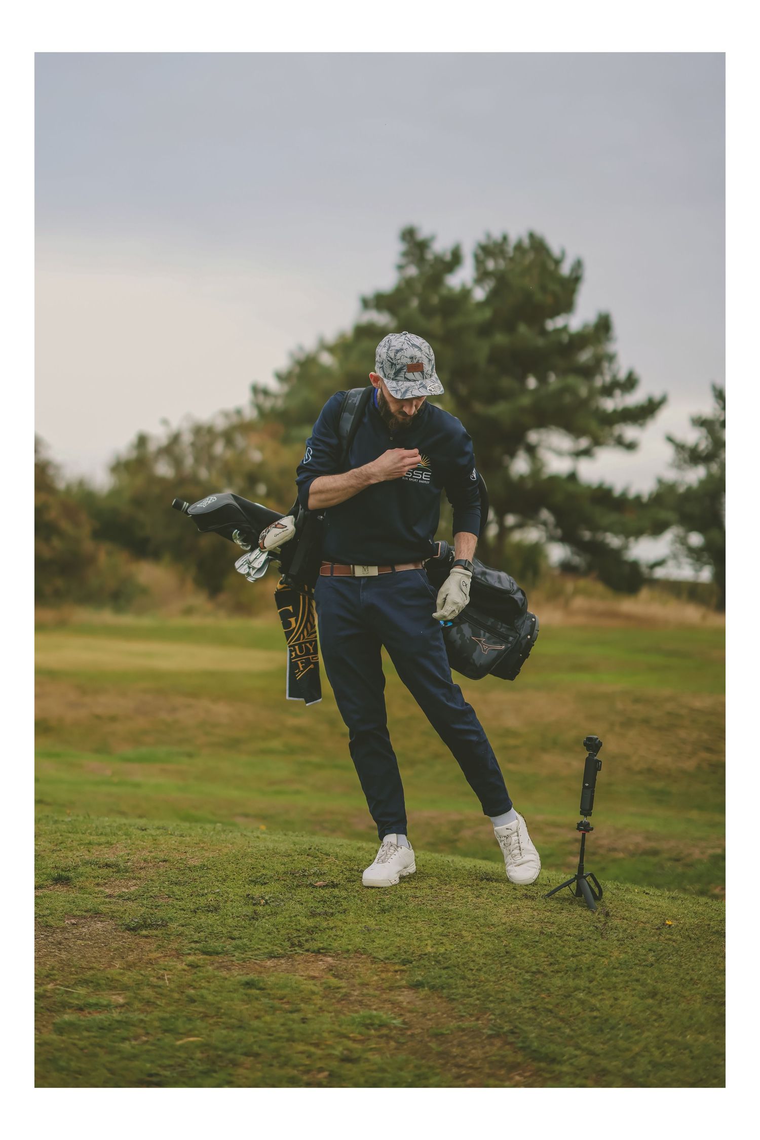 Male golfer carrying bag and equipment while walking across a golf course on an overcast day.