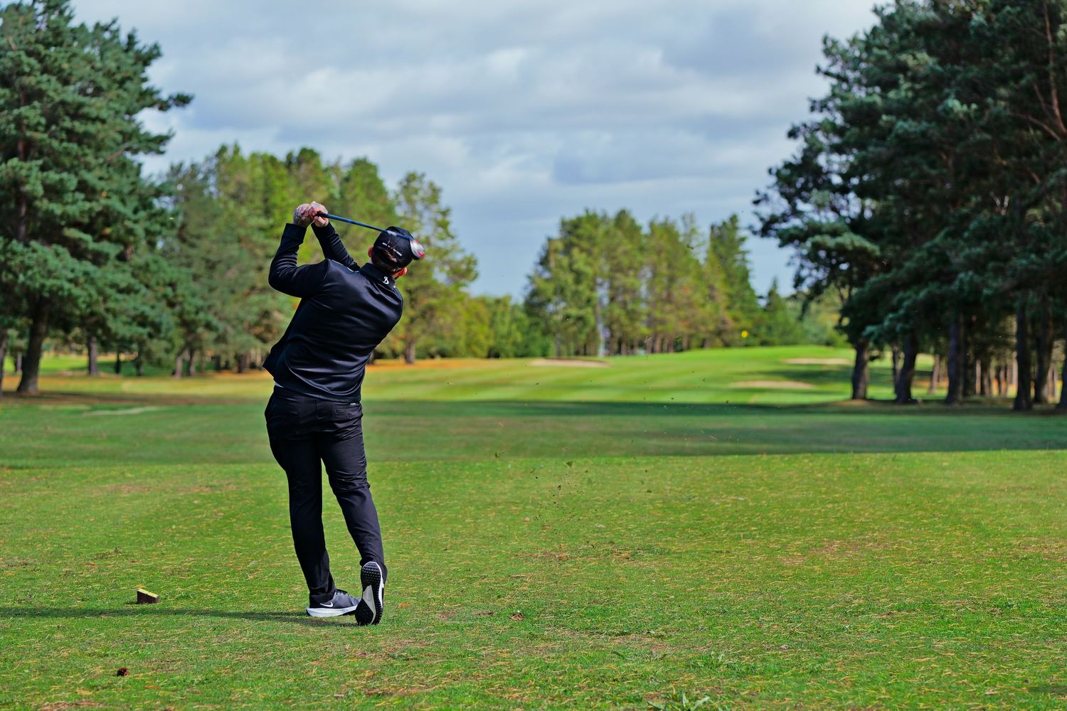Golfer in black outfit completing a swing on a lush green golf course surrounded by tall pine trees.