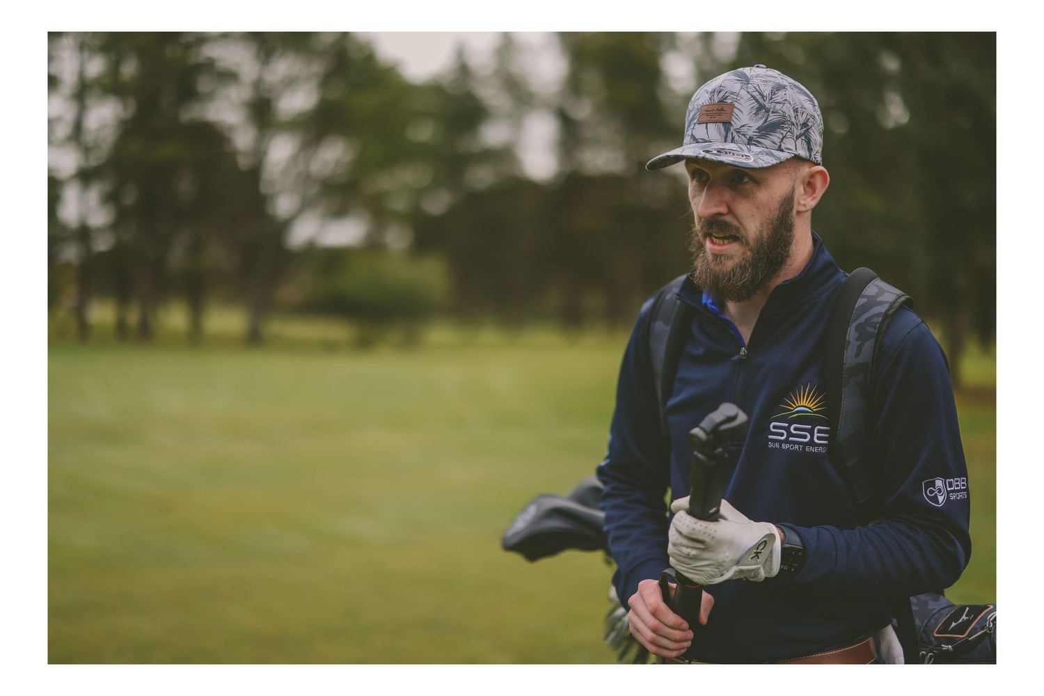 Bearded male golfer wearing a cap and navy vest carrying golf clubs on a green course.