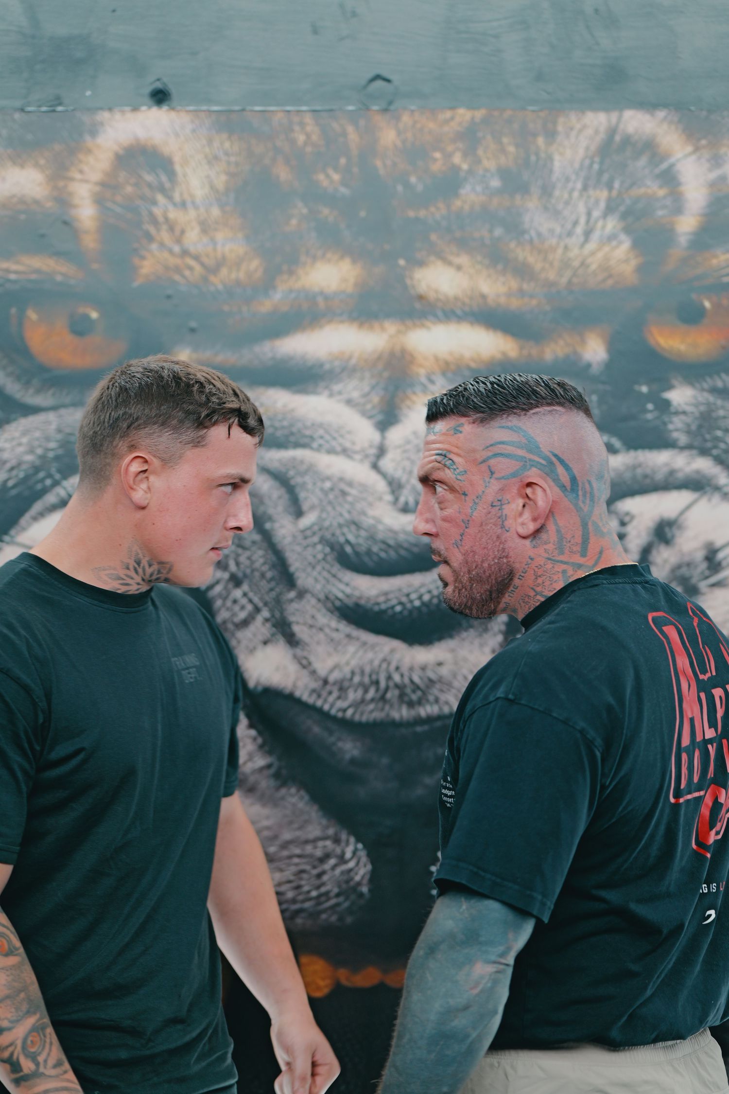 Two heavily tattooed men face each other in front of a large tiger graffiti mural outdoors.