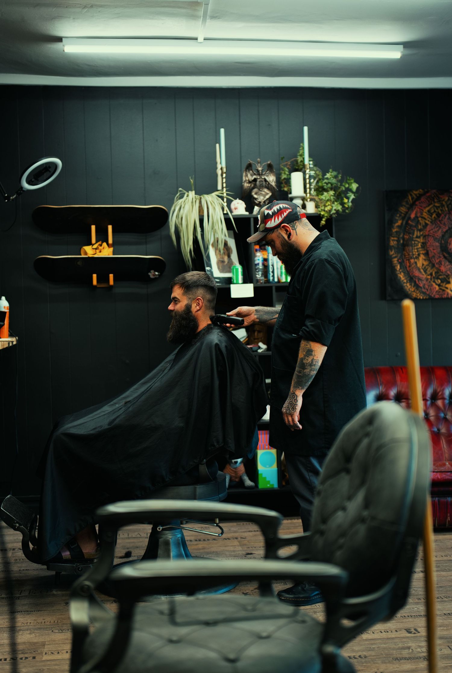 A tattooed barber cutting a bearded client's hair in a stylish dark-walled barbershop with eclectic decor.
