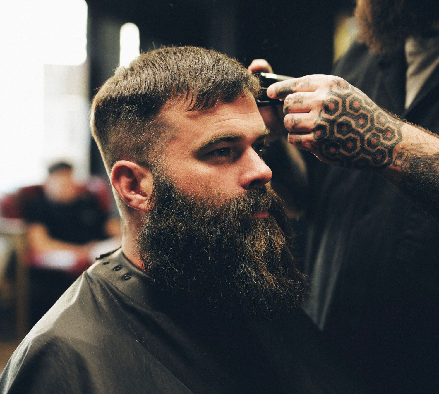 Man with a large beard getting a haircut from a tattooed barber in a barbershop.