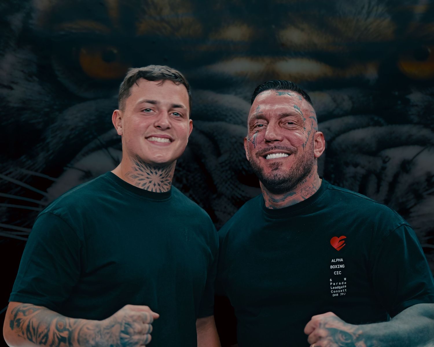 Two smiling tattooed men posing together wearing dark green shirts in front of a tiger mural.