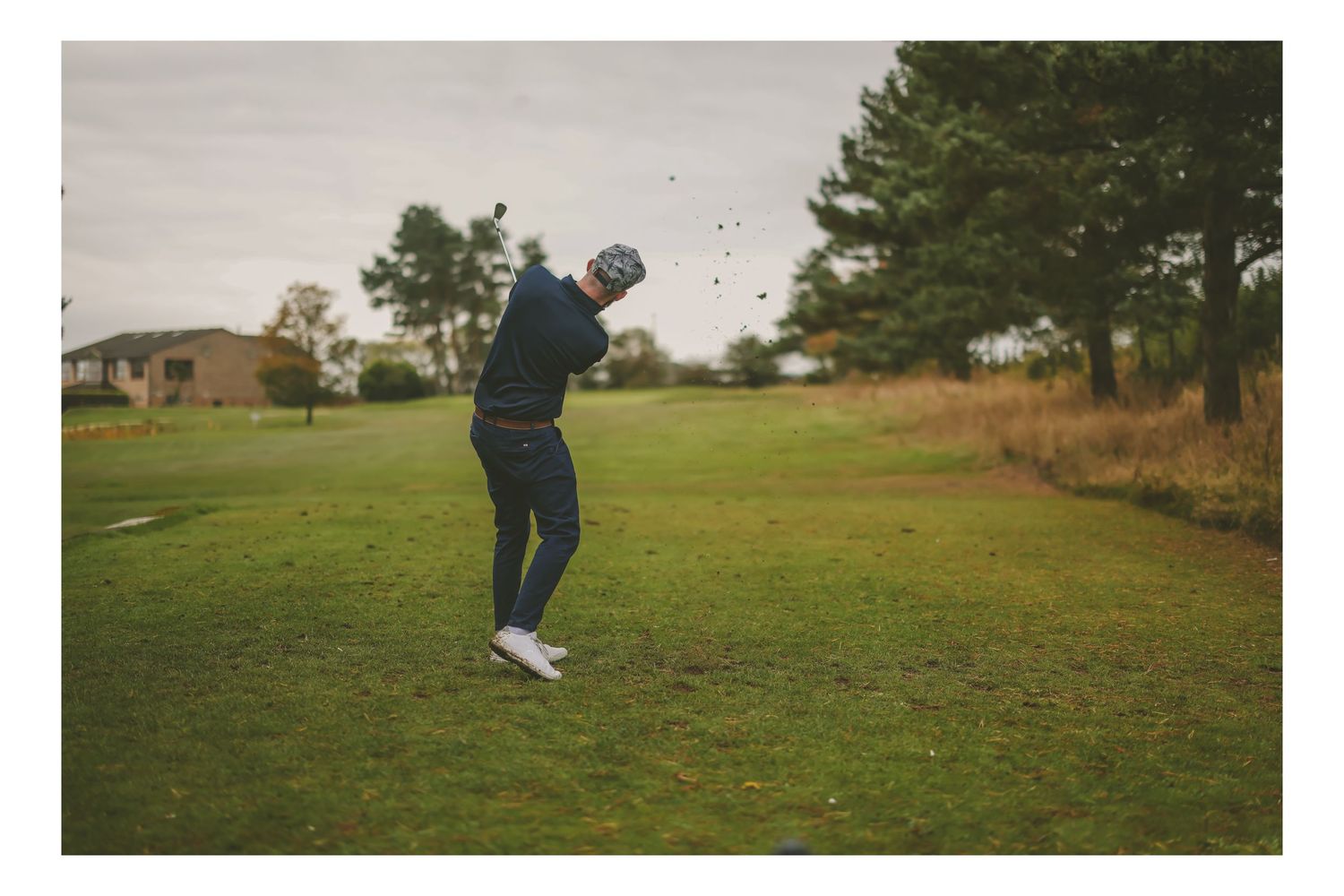 A golfer swings an iron shot on a green fairway surrounded by trees on a cloudy day.