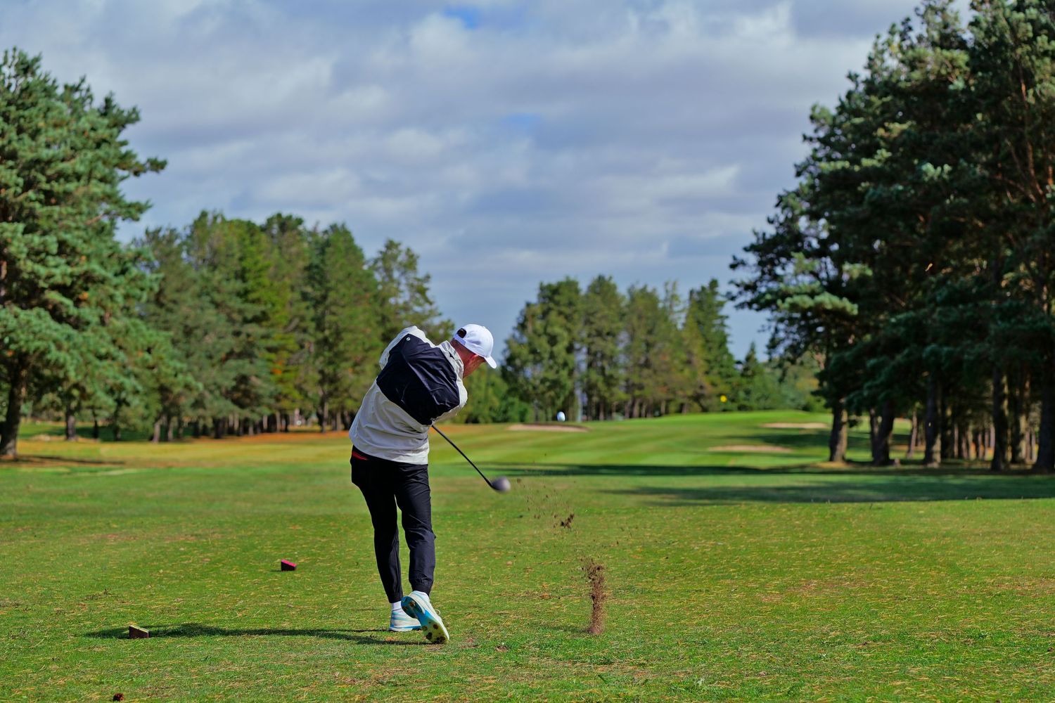 Golfer in white cap and jacket swinging a club on a well-maintained golf course with pine tree backdrop.