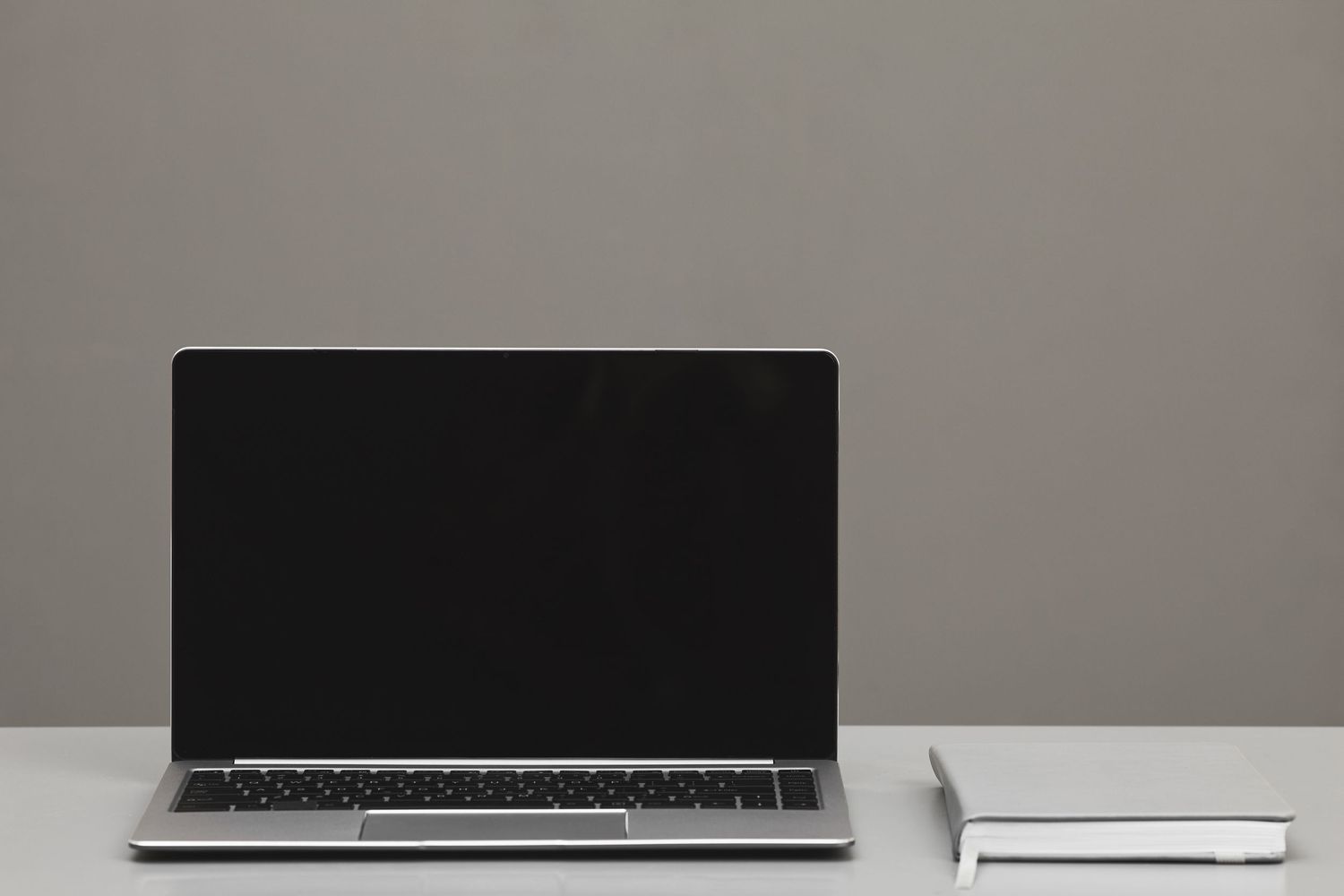 Open laptop with blank screen beside a small book on a clean white desk against a gray wall.
