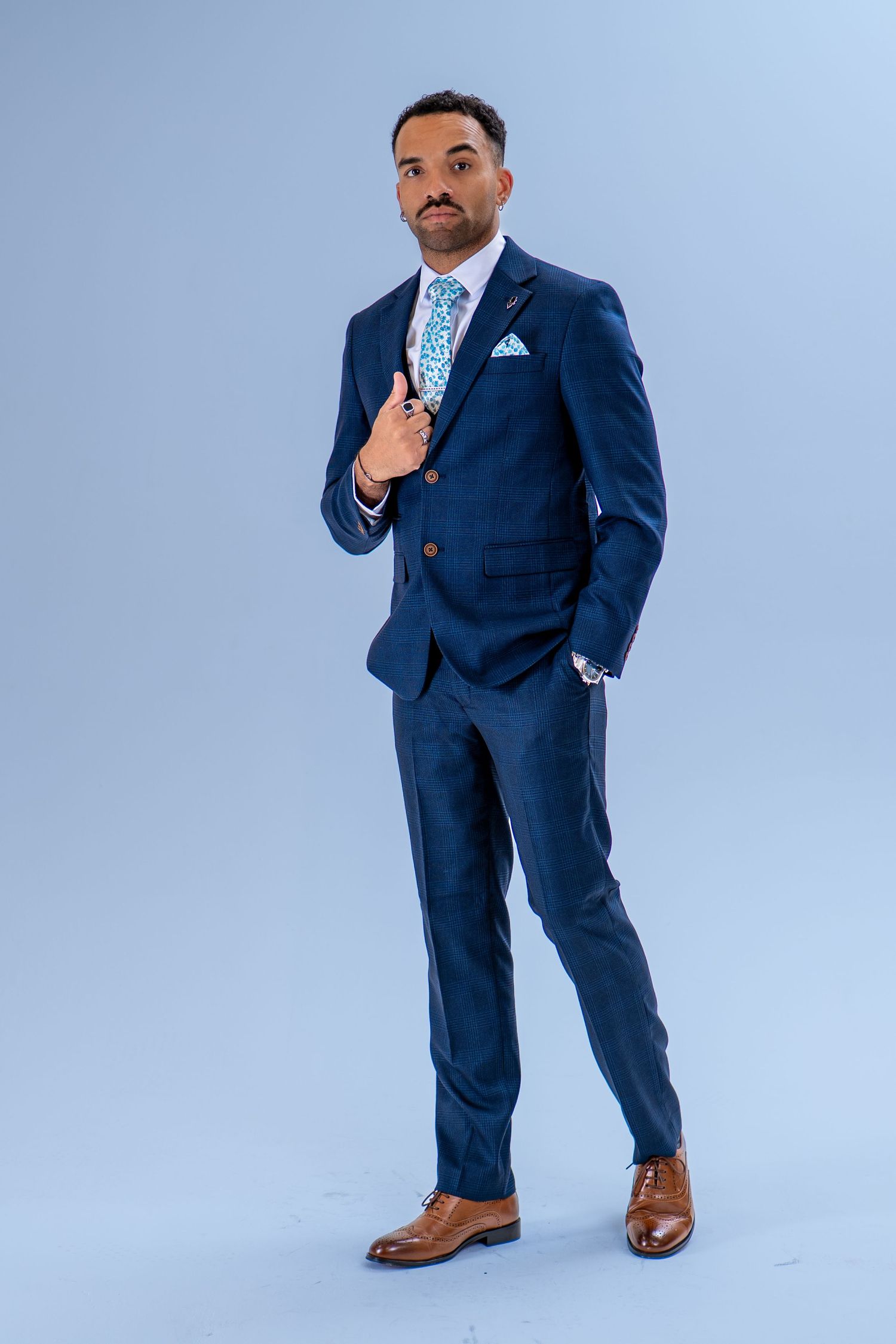Man in navy blue suit with floral tie, pocket square, and brown oxford shoes posing against light blue background.