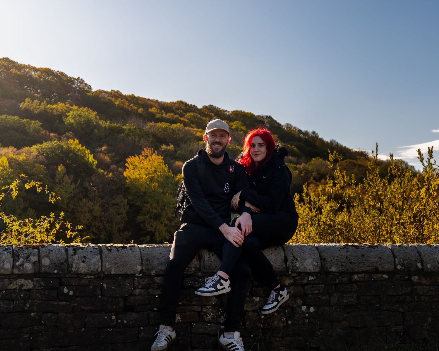 Smiling couple in dark clothing sits together on a stone wall with a hillside of golden autumn trees and blue sky behind them.