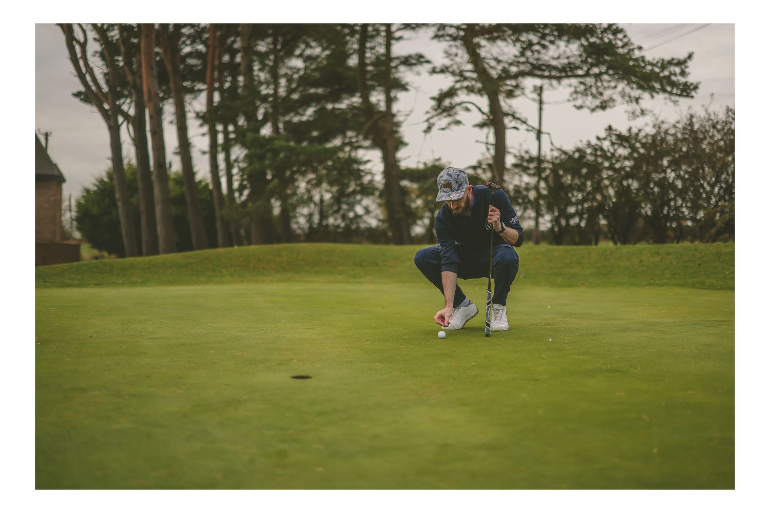 A golfer crouches on a putting green carefully lining up a putt on a cloudy day.