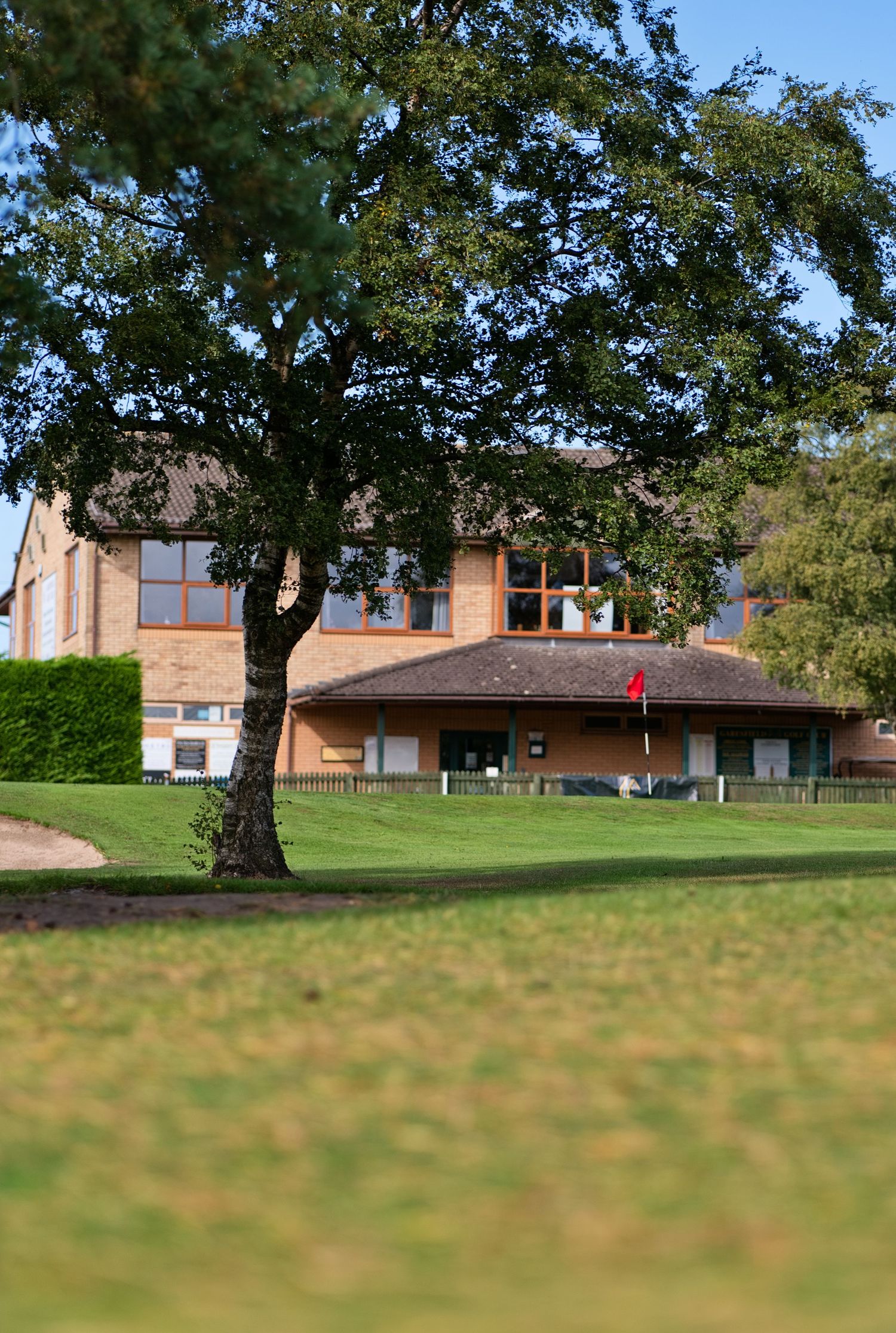 Brick clubhouse with large windows and a red flag sits beyond a mature tree and manicured green lawn on a sunny golf course.