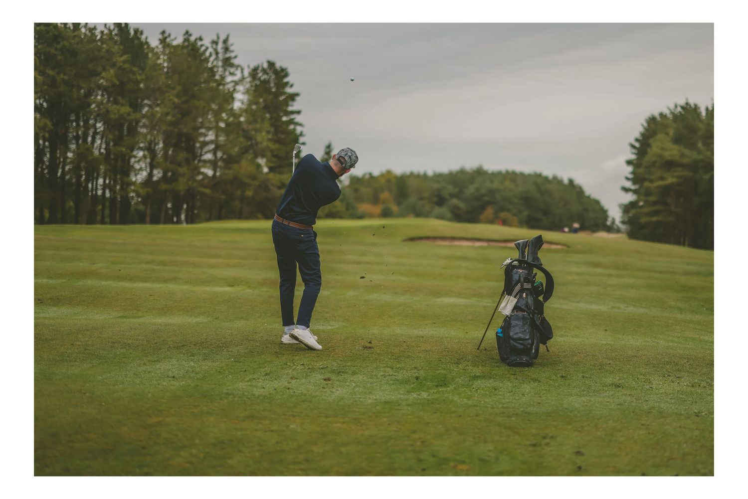 A golfer mid-swing on a lush fairway with a golf bag nearby, surrounded by tall trees.