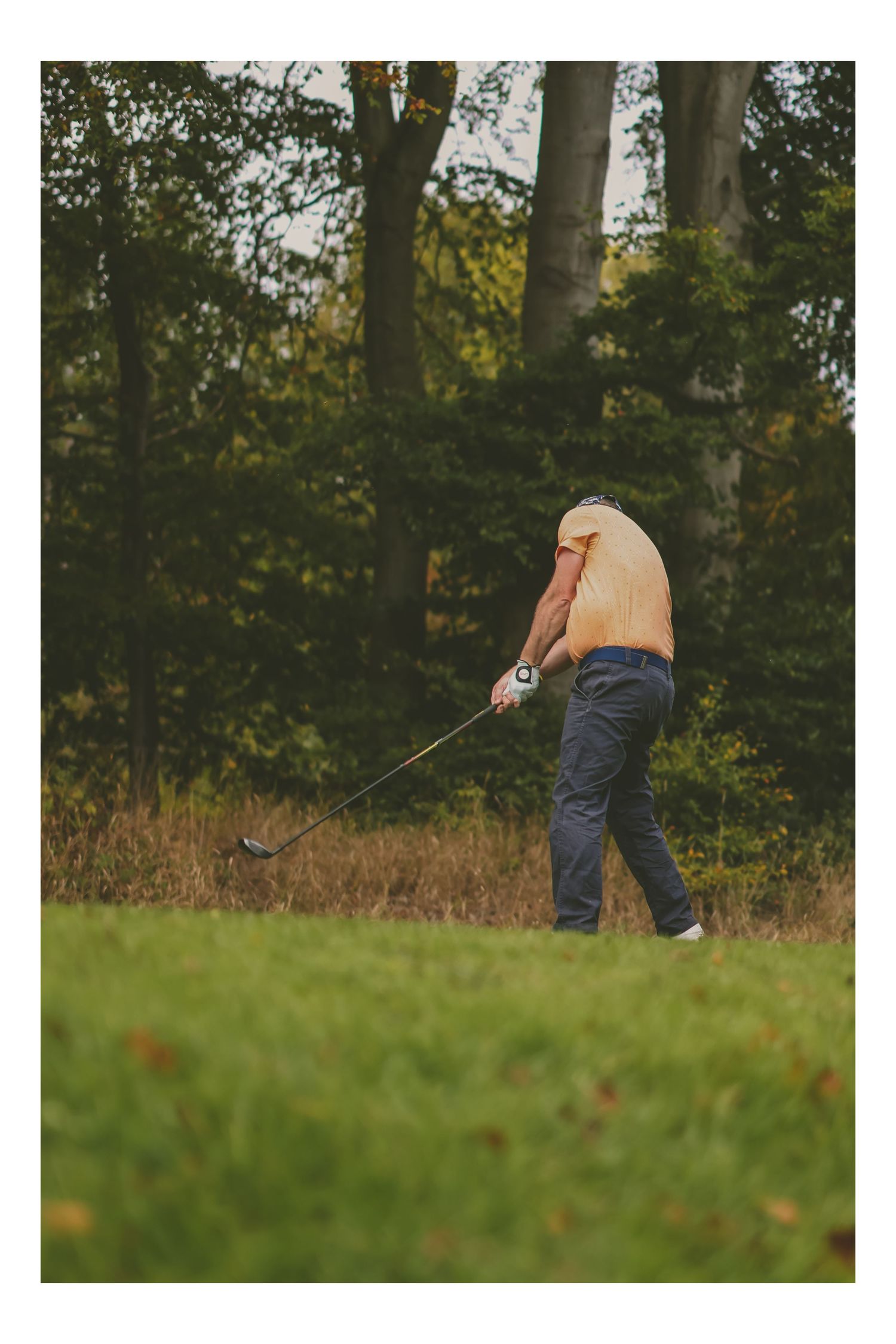 A golfer in a tan shirt and grey trousers plays a shot from the rough near woodland trees.
