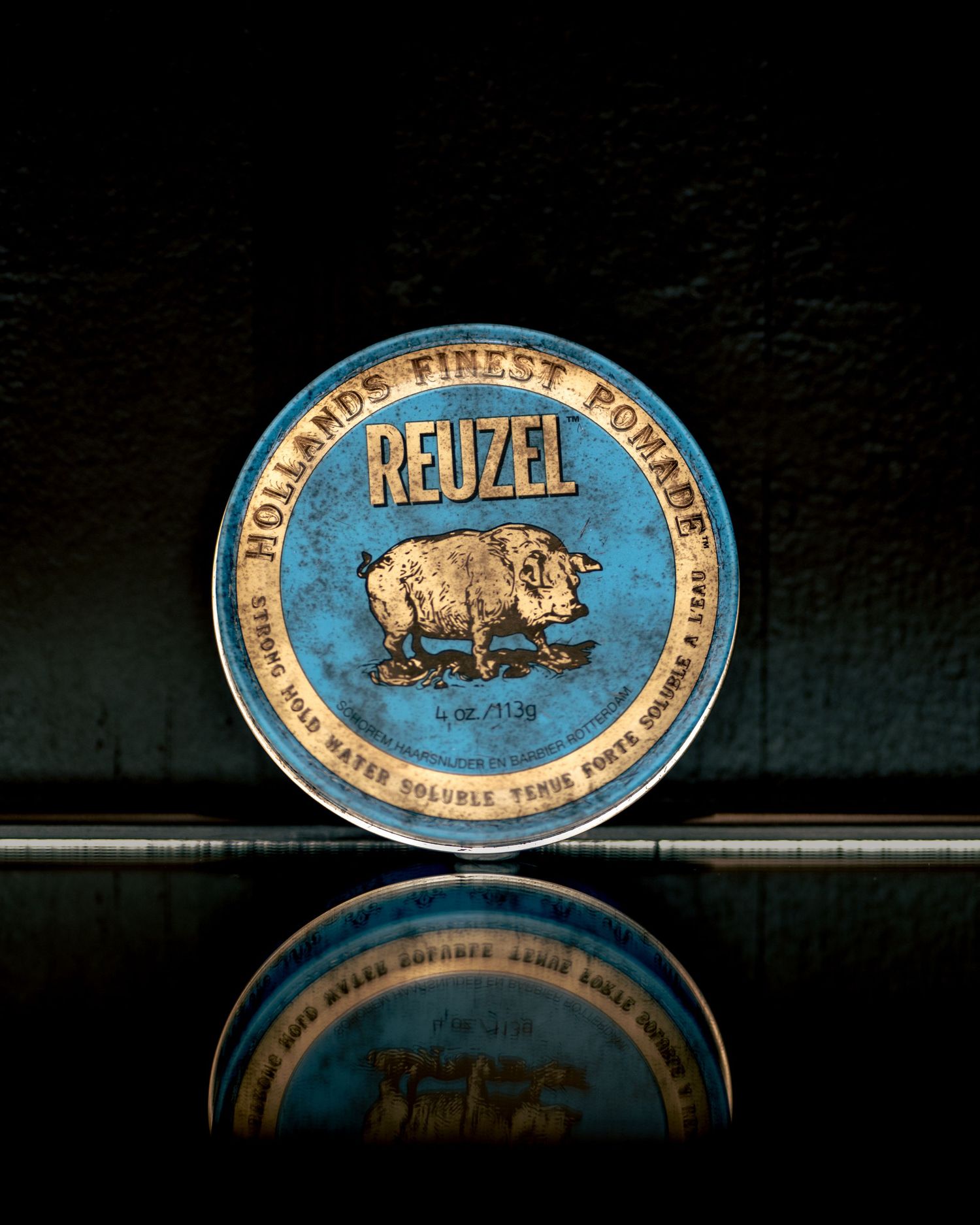 Reuzel blue strong hold water soluble hair pomade tin on a dark reflective surface.