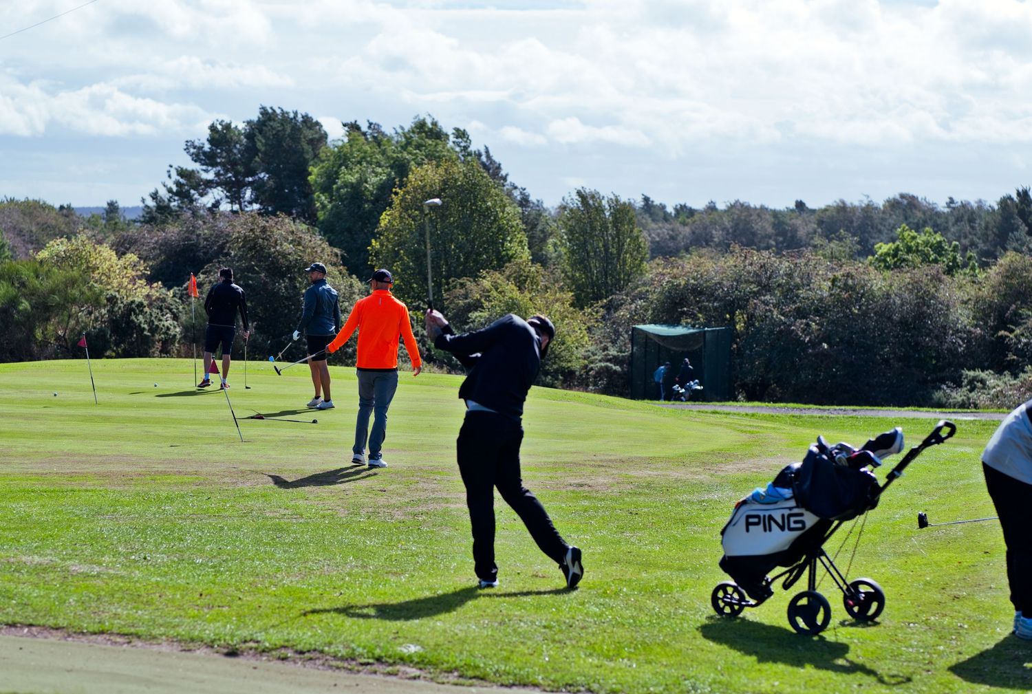 Golfer mid-swing with a Ping trolley on a sunny golf course with other players nearby.