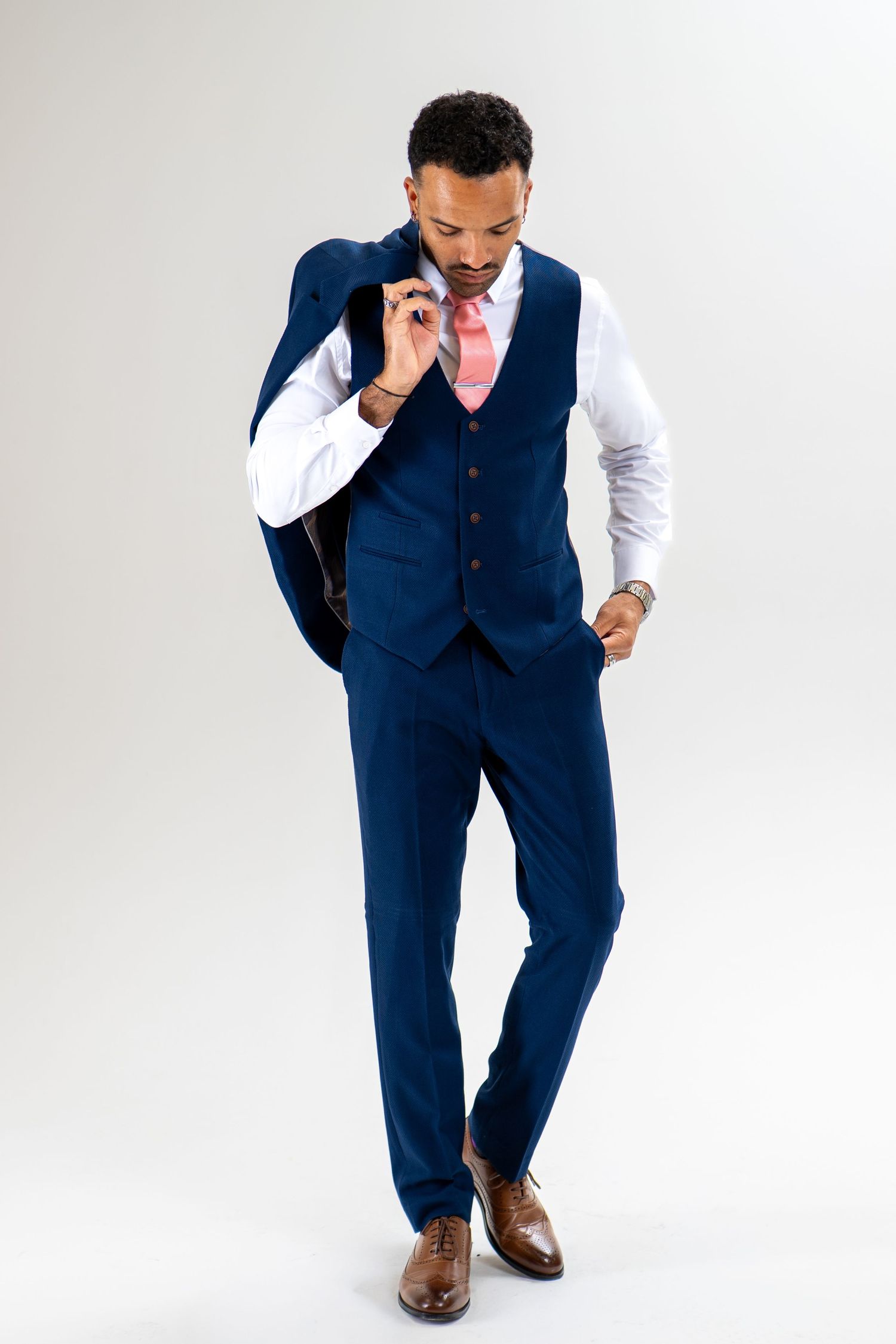 Man in a navy blue three-piece suit with pink tie and brown leather shoes, holding his jacket over his shoulder against a plain backdrop.