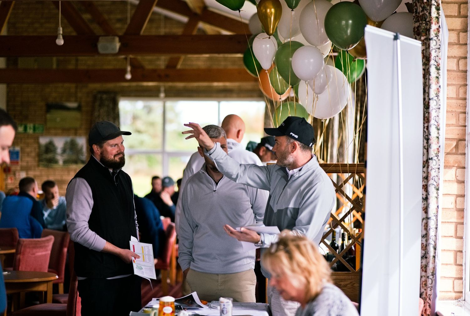 Group of men in conversation at a rustic event venue decorated with green, white and gold balloons, with guests seated at tables nearby.