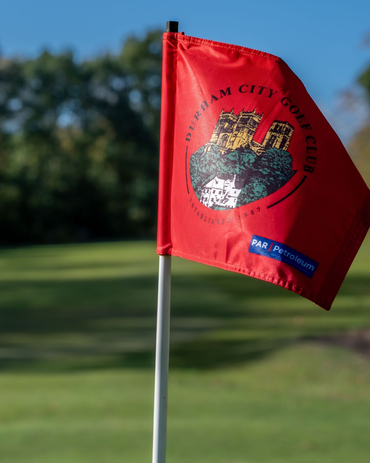 Red Durham City Golf Club flag with castle crest logo flying on a white pole over a sunlit green fairway with trees behind.