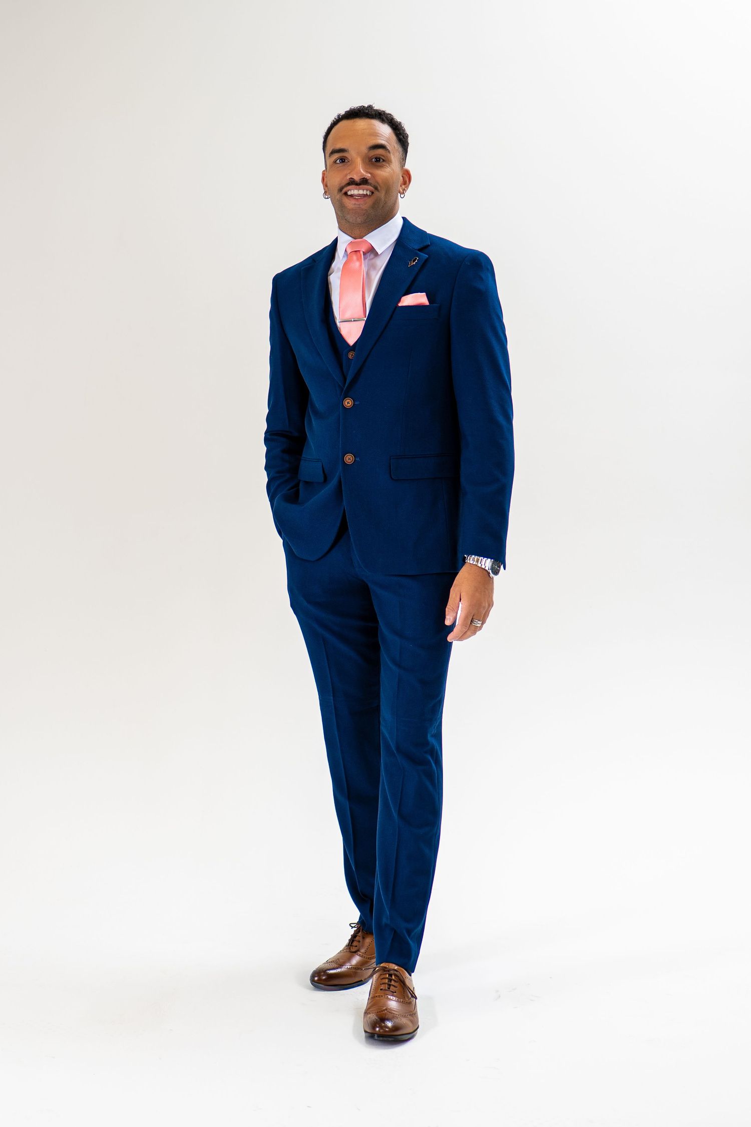Man in navy blue three-piece suit with pink tie and pocket square, brown dress shoes, smiling on white background.
