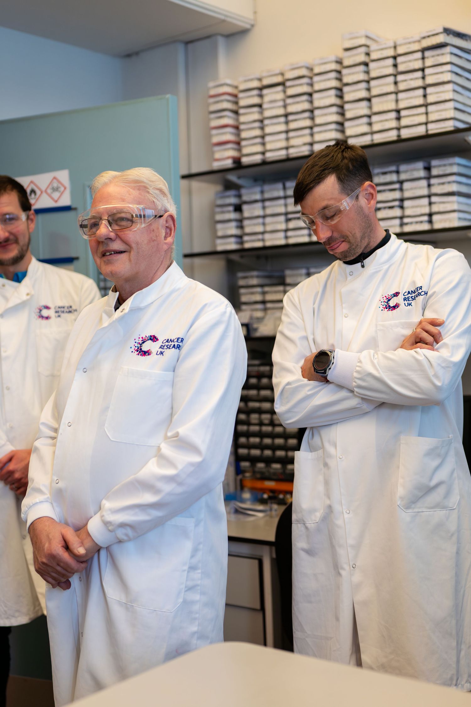 Three researchers in white Cancer Research lab coats stand smiling in a laboratory with shelves of stacked sample plates behind them.