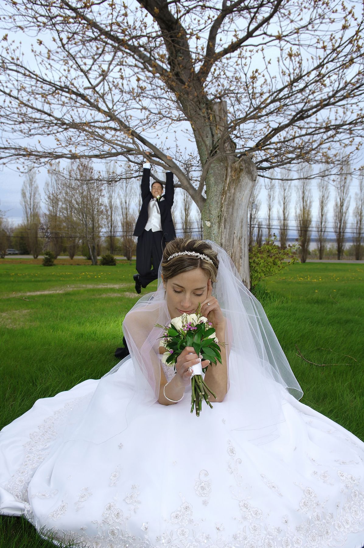 Bride sitting on green lawn holding her bouquet with groom in background
