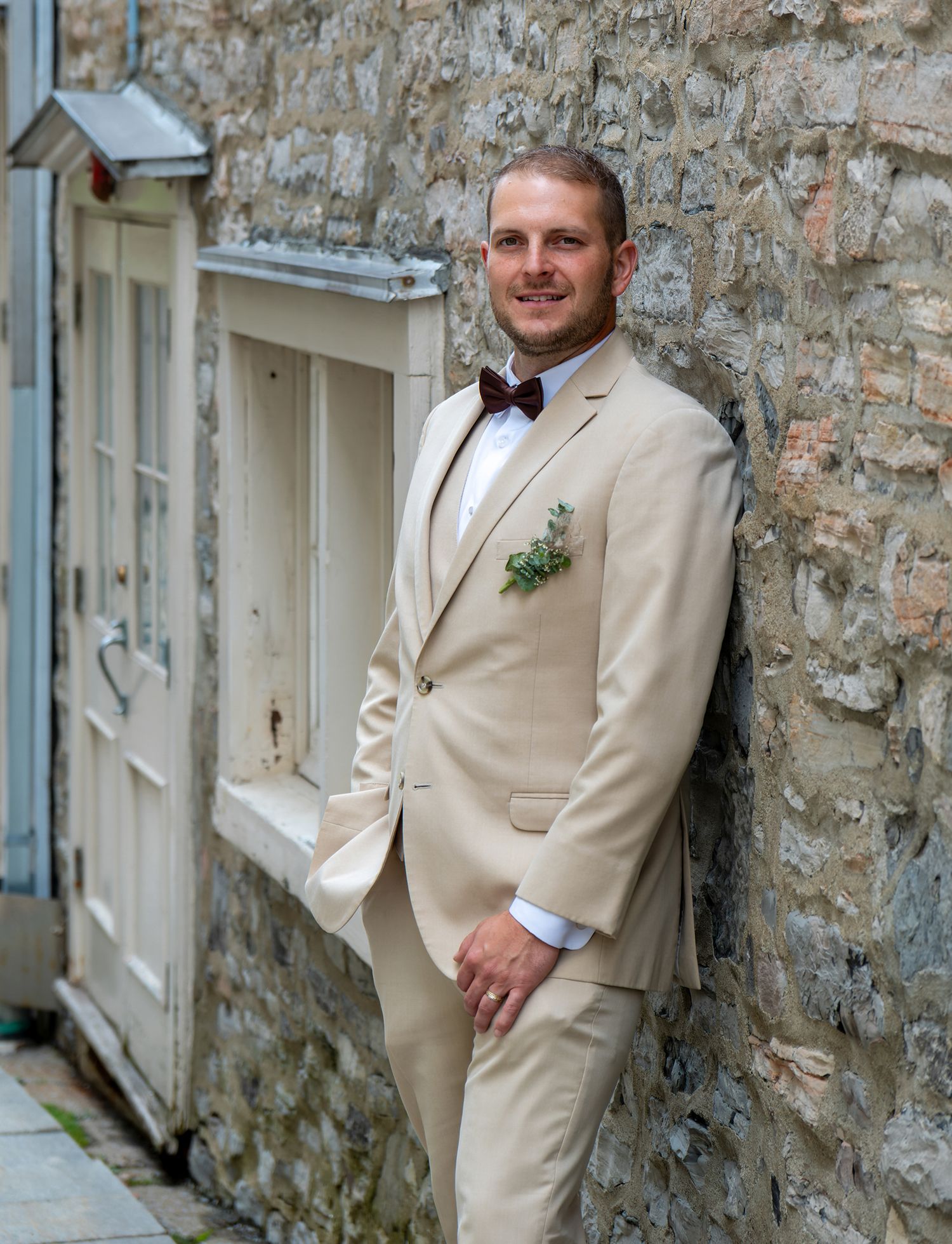 Portrait of a groom in a tan suit leaning against a historic stone wall in the streets of Old Quebec