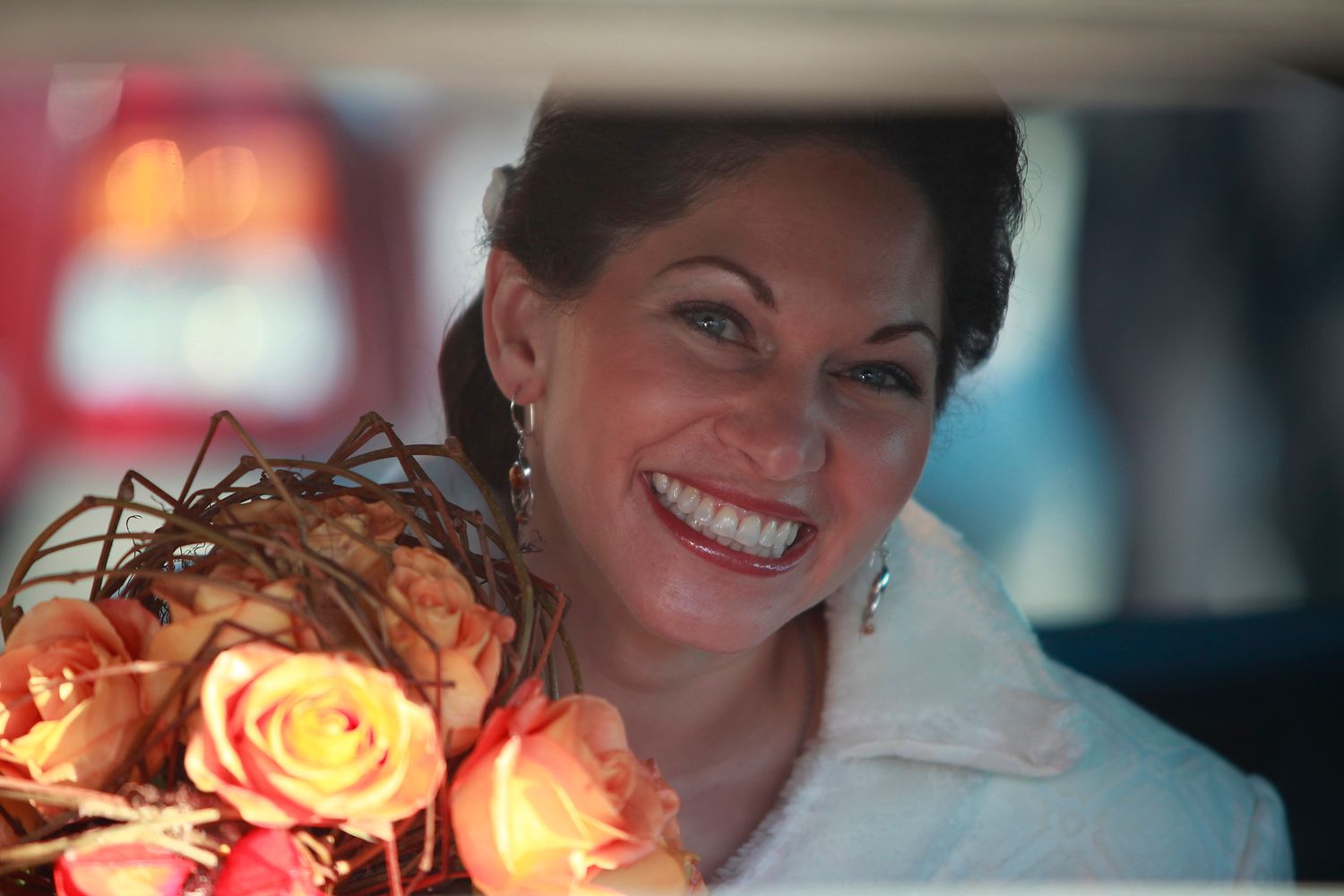 Bride smiling from a wedding car window holding a warm orange rose bouquet