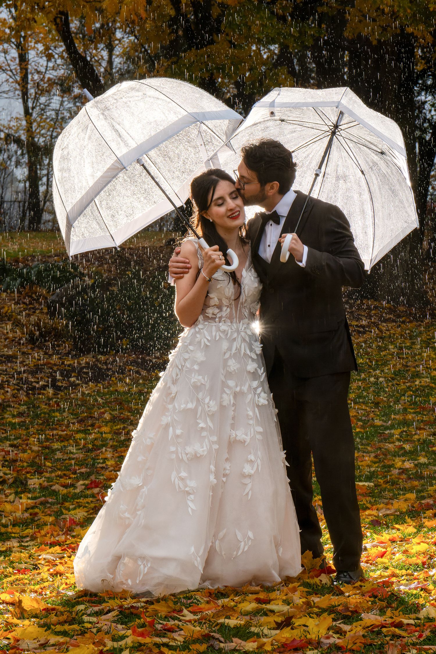 A happy bride and groom kiss under a clear umbrella during an autumn rain in a Quebec City park