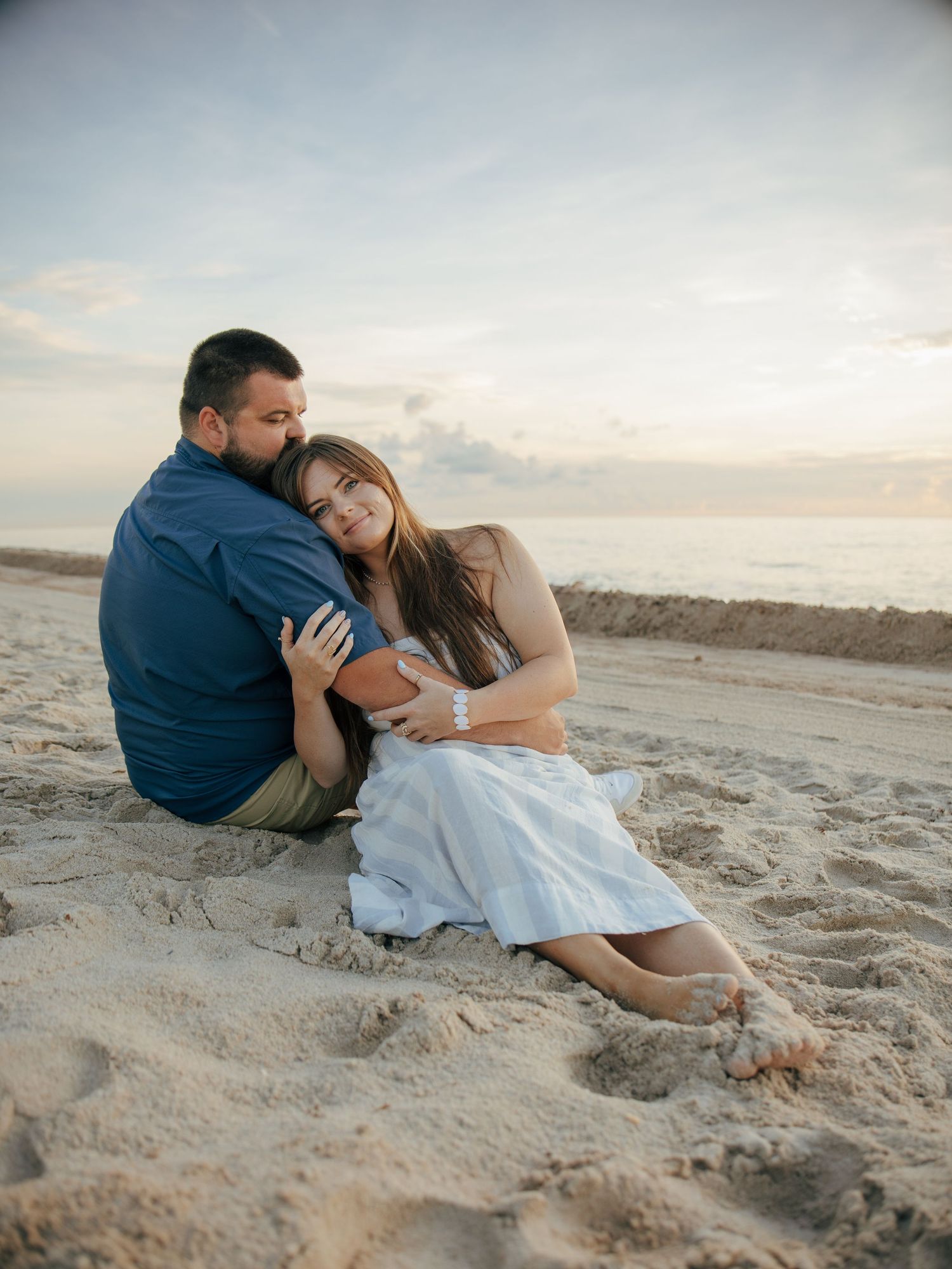 Couple sitting together on sandy beach at sunset, woman embracing man from behind in tender maternity photo session.