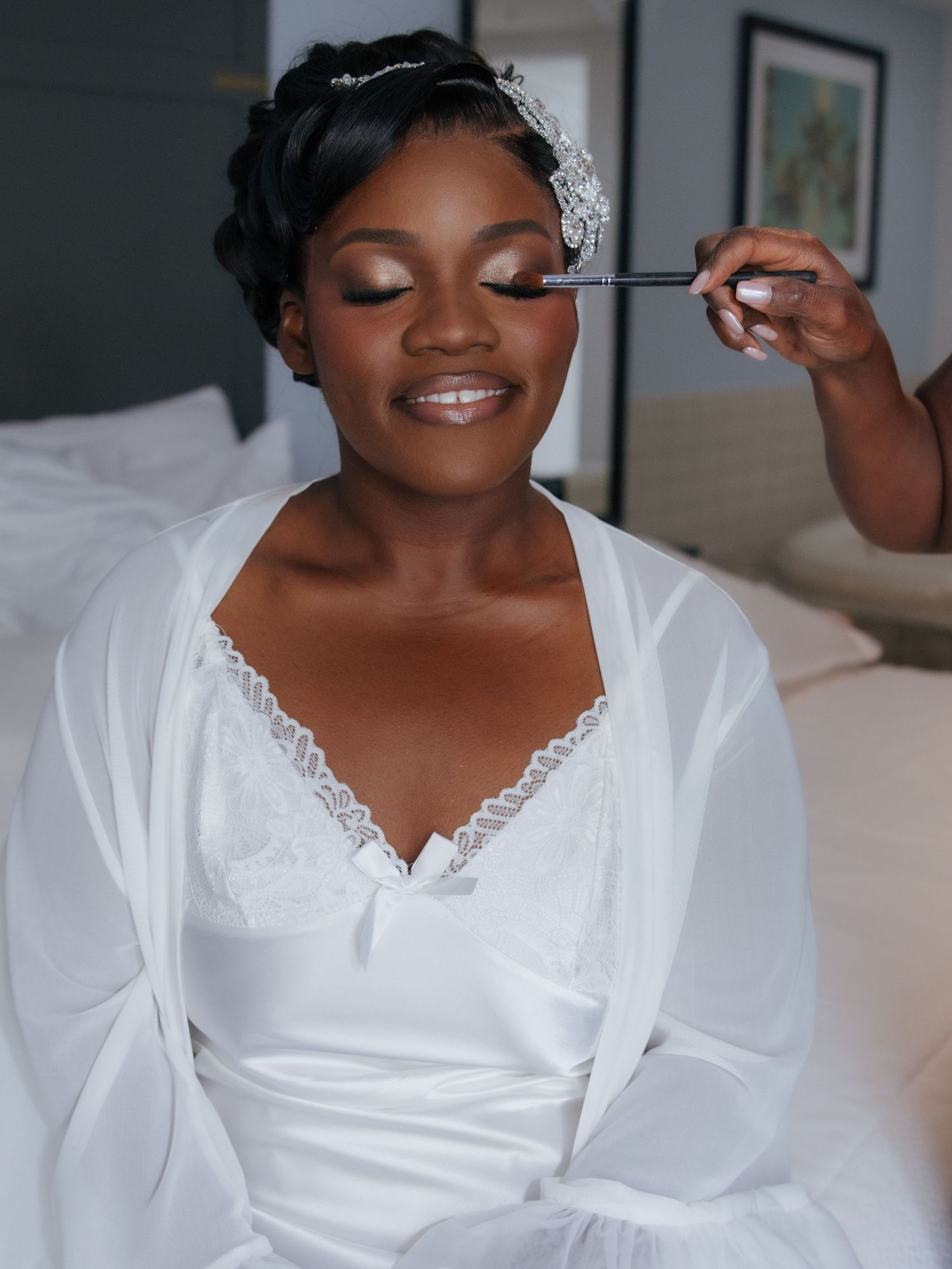A bride gets her makeup applied while wearing a white lace robe and floral hair accessory on her wedding day.