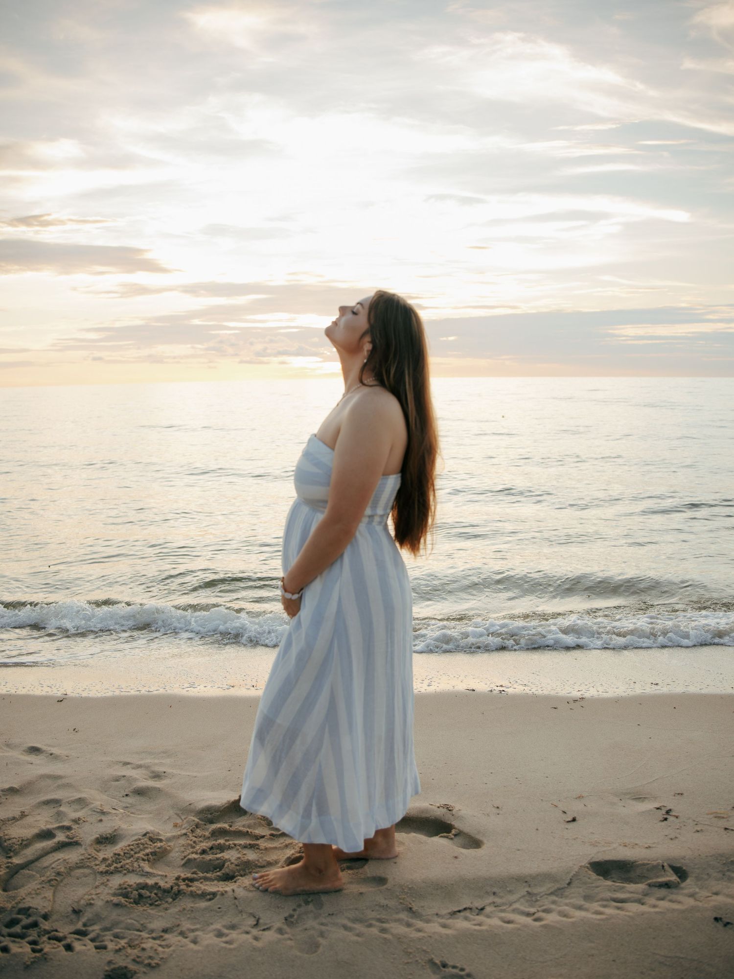 Pregnant woman in blue striped strapless dress standing on beach gazing upward at golden sunset sky.