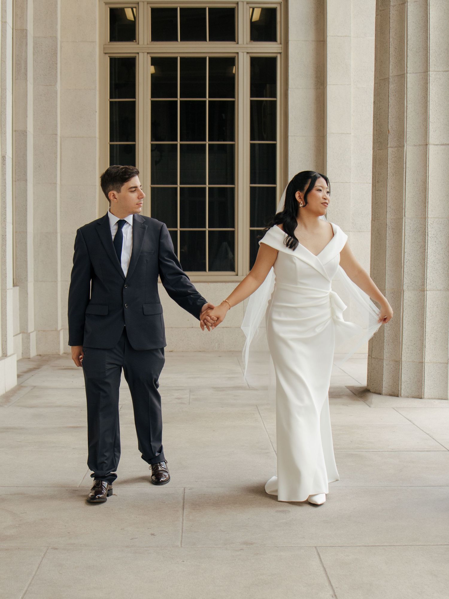 Bride in sleek white gown and groom in navy suit walk hand in hand between stone columns.