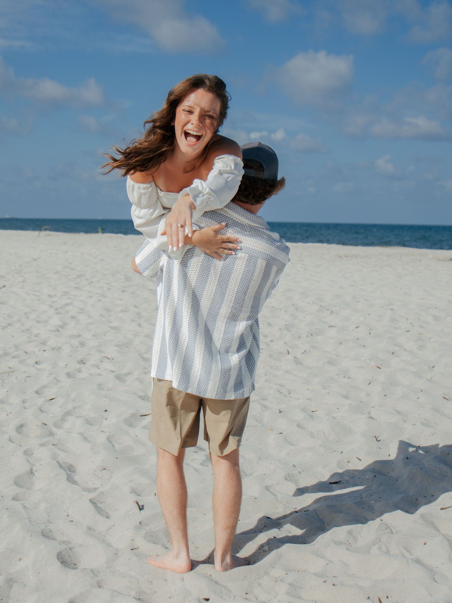 Joyful woman in white outfit laughing while being lifted in a hug on a bright sandy beach.