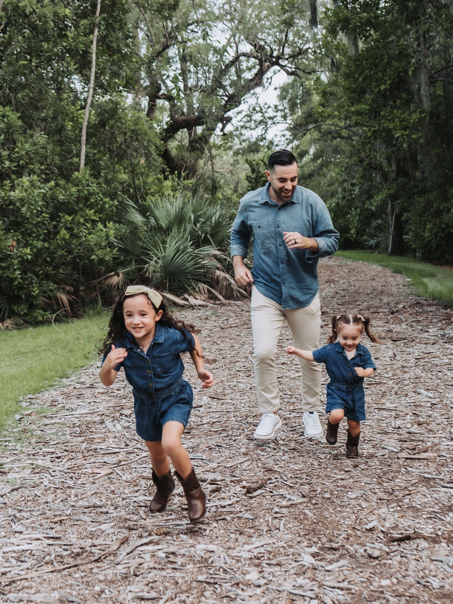 A father joyfully runs with his two young daughters along a tree-lined mulch path outdoors.