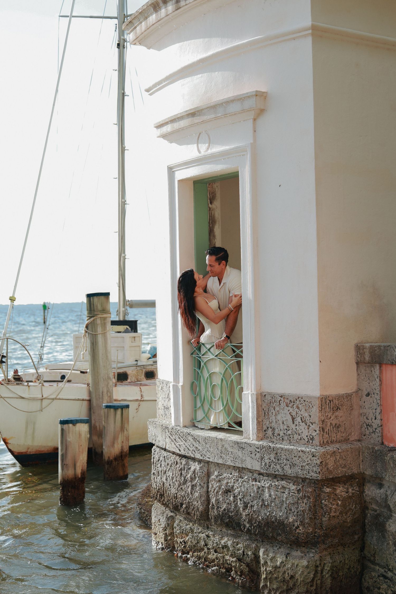 A romantic couple embraces in a waterfront doorway with a sailboat docked beside a historic building.