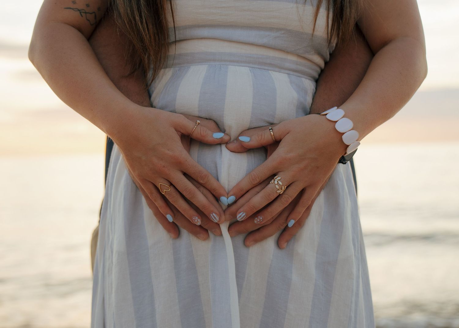 Pregnant woman forming heart shape with hands on baby bump in striped dress at beach sunset.