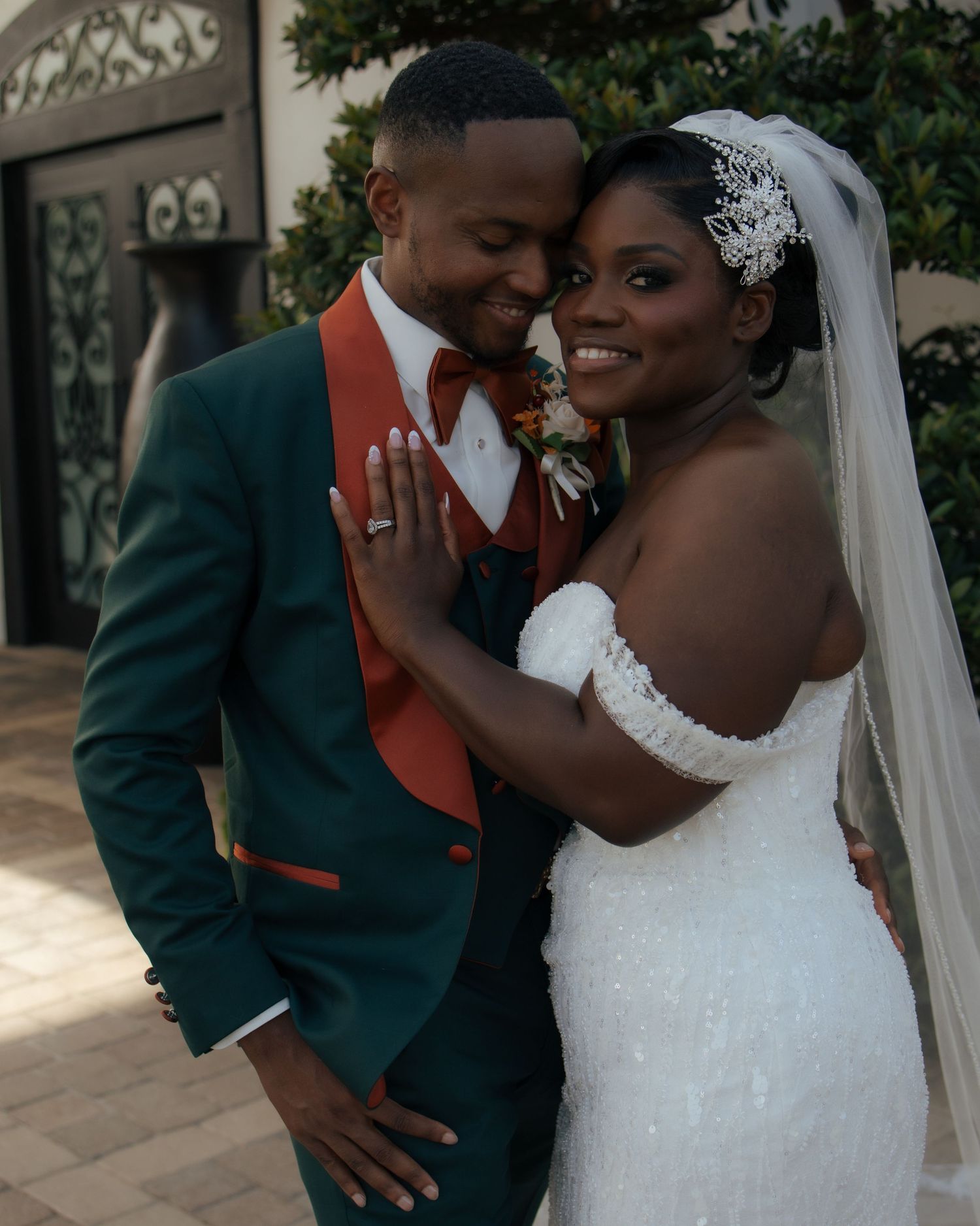 A smiling bride in a beaded gown poses with her groom in a green and orange tuxedo outdoors.