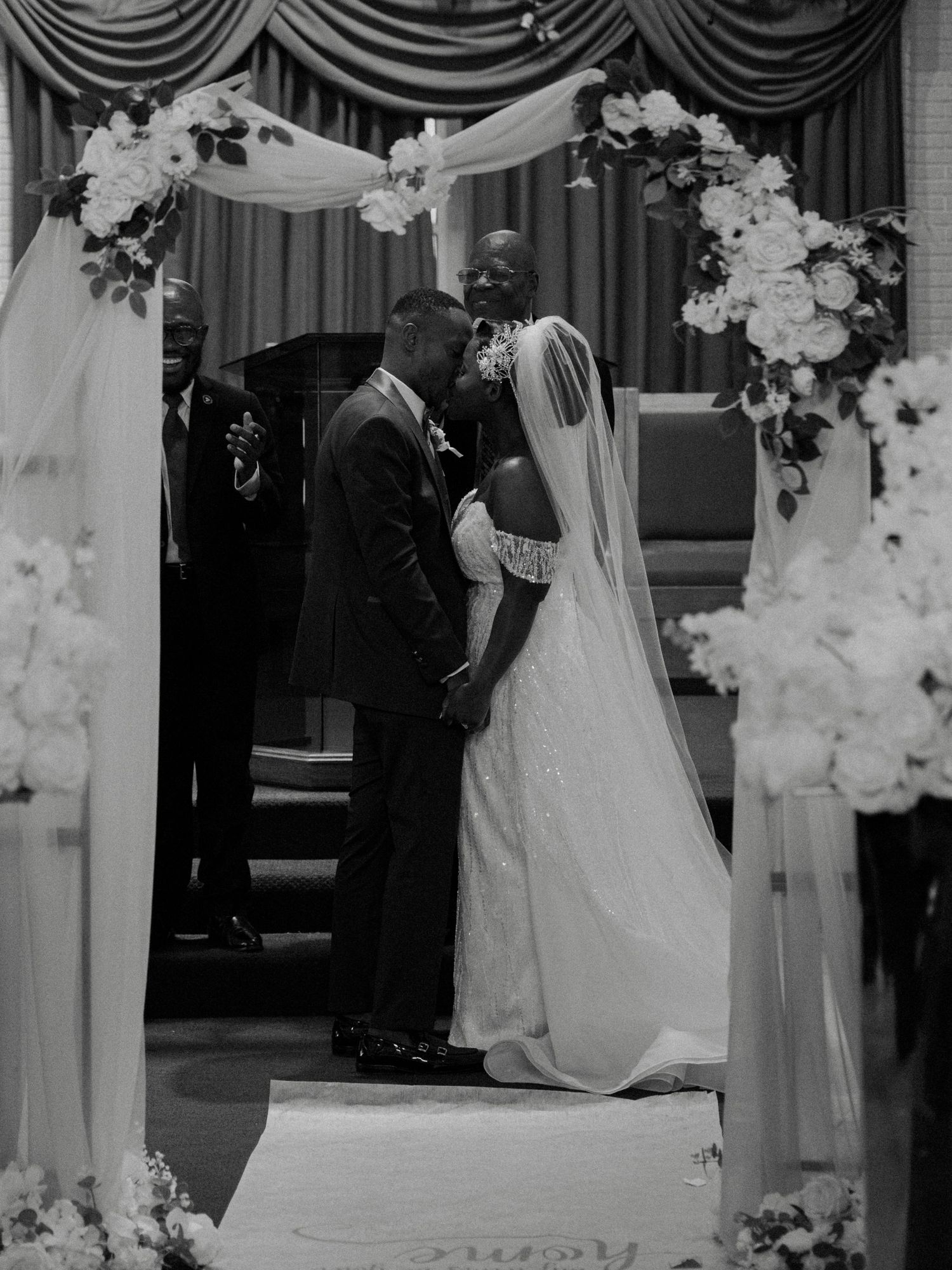 Black and white photo of a bride and groom sharing their first kiss under a floral arch at the altar.