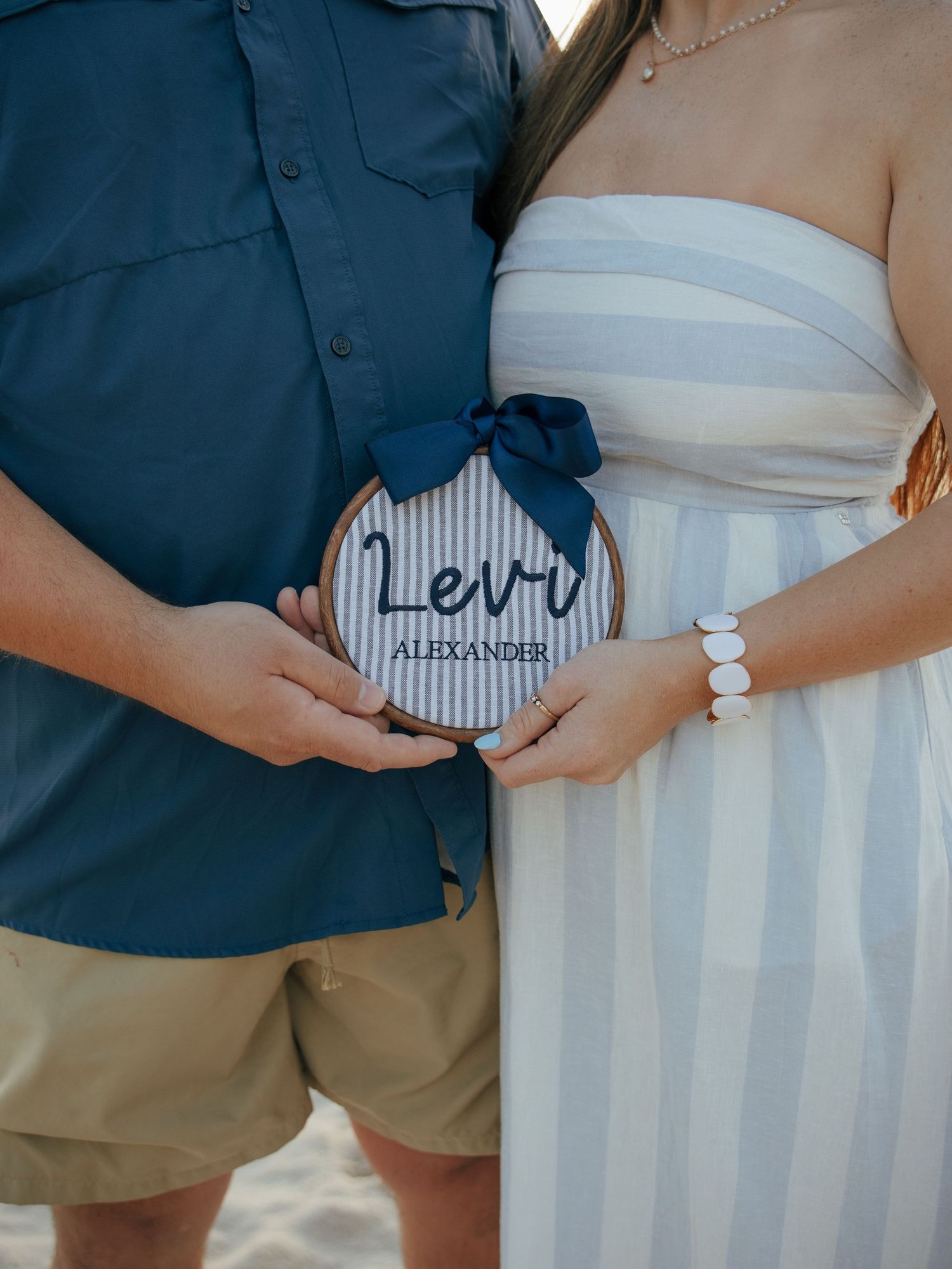 Couple holding round wooden sign reading Levi Alexander at beach maternity photo session.