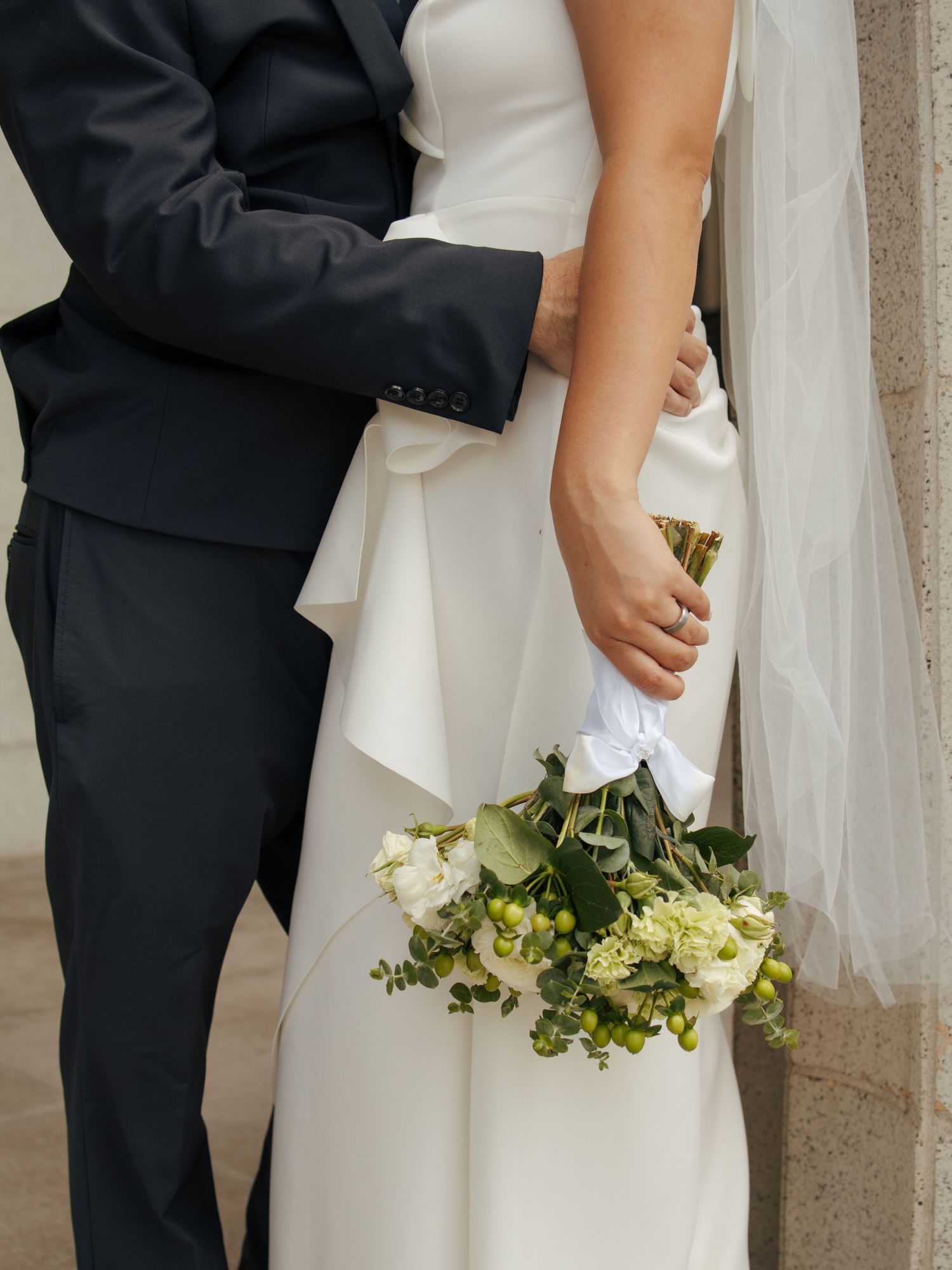 Bride and groom embrace against stone wall, bride holding white and green wedding bouquet.
