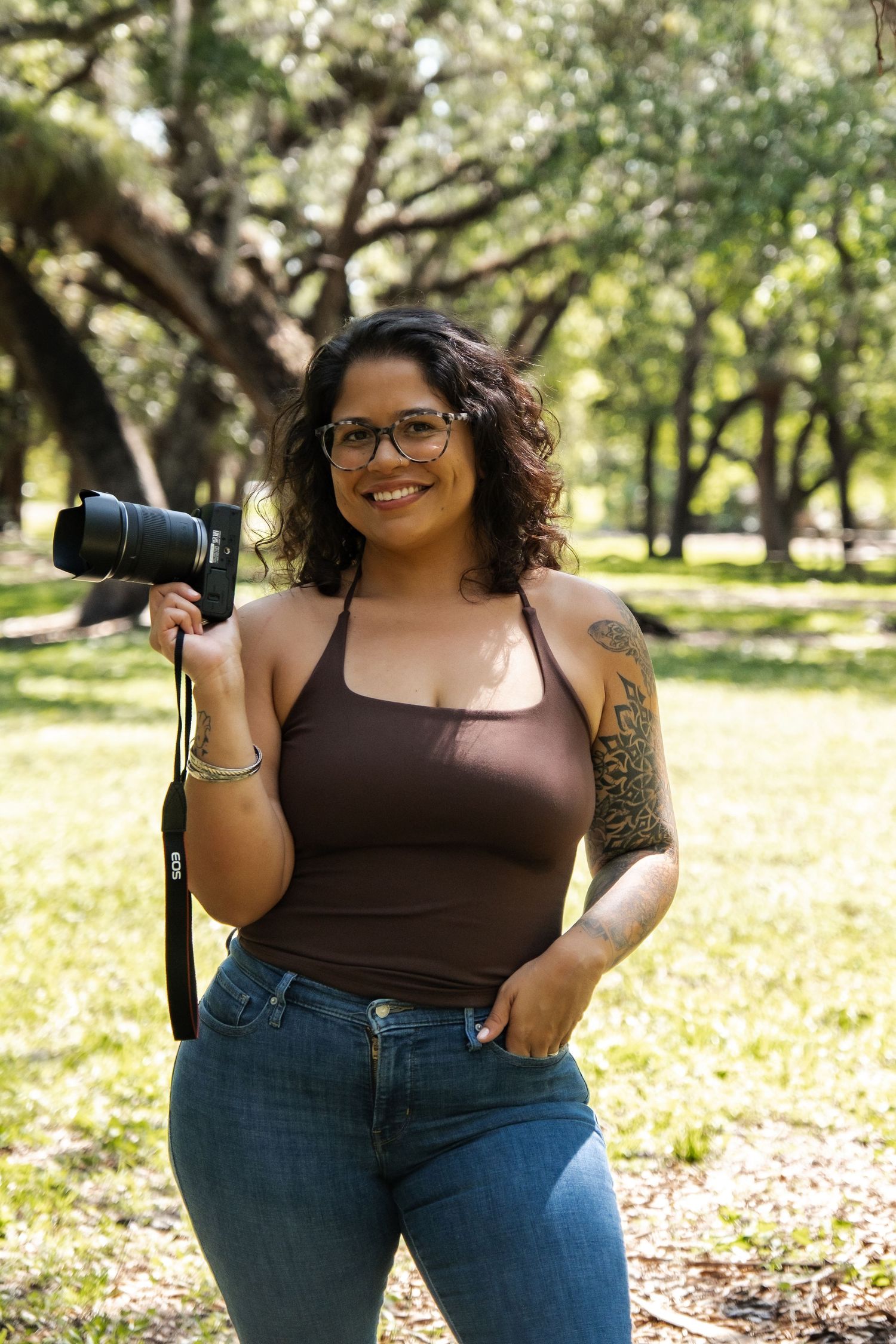 Female photographer smiling in park holding professional camera, wearing glasses and brown top.