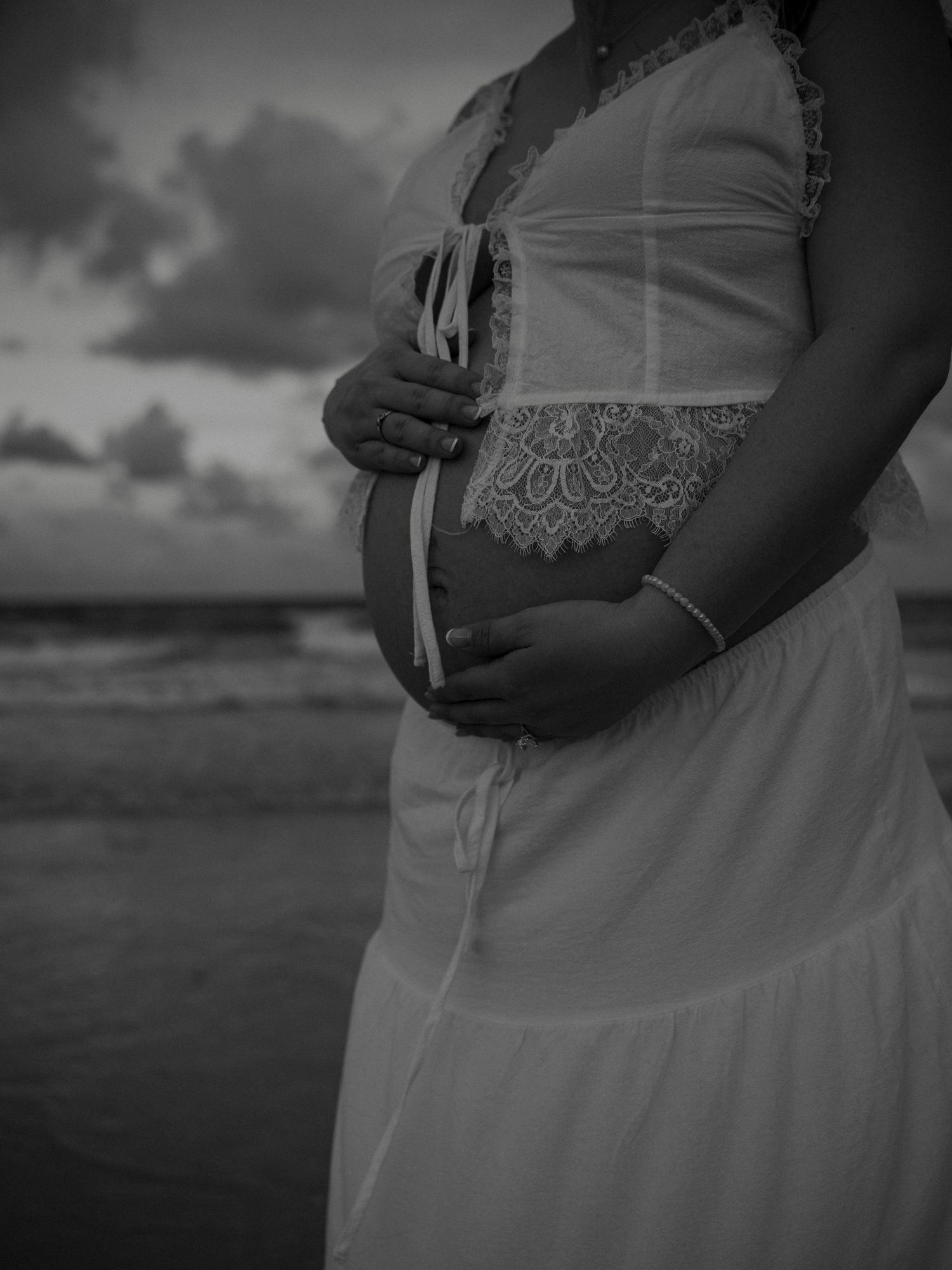 Black and white close-up of pregnant woman in white lace dress cradling baby bump on beach.