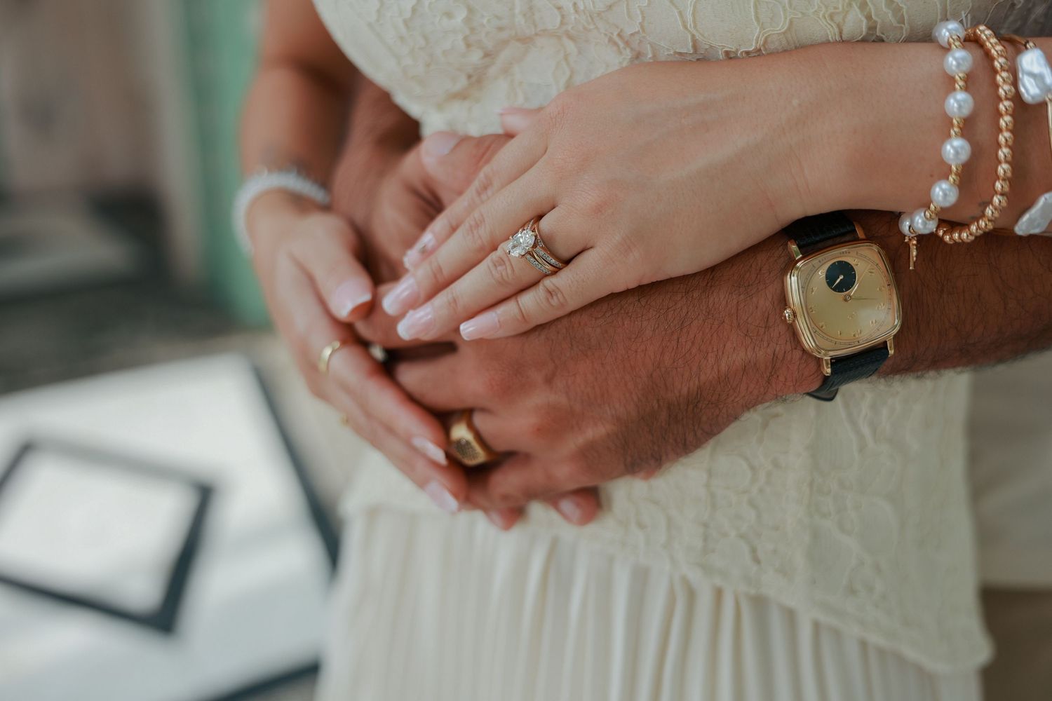Close-up of a couple\u2019s hands showing an engagement ring, gold band, pearl bracelet, and smart watch.
