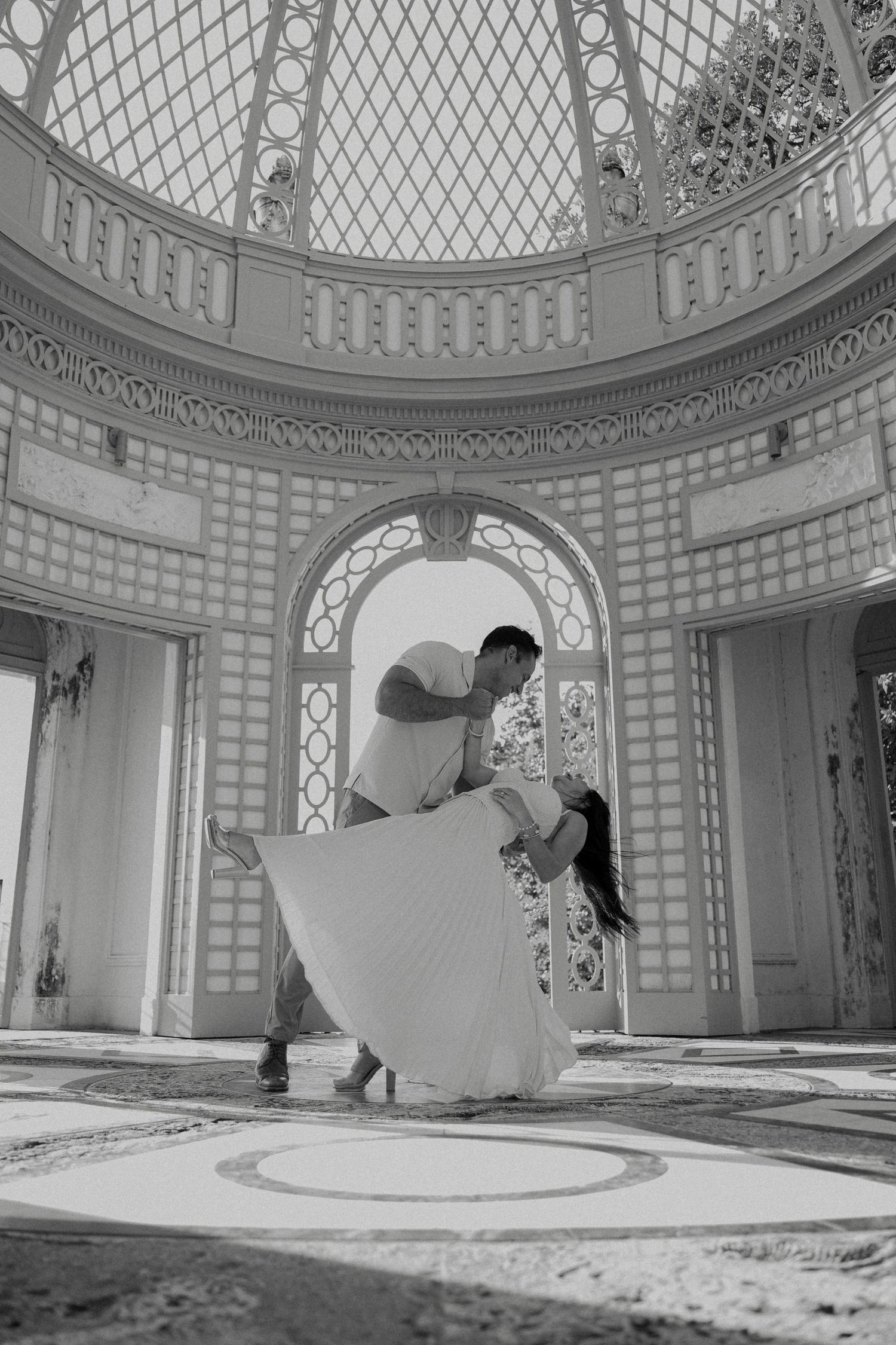 A couple dances joyfully in black and white inside an elegant domed hall with decorative archways.