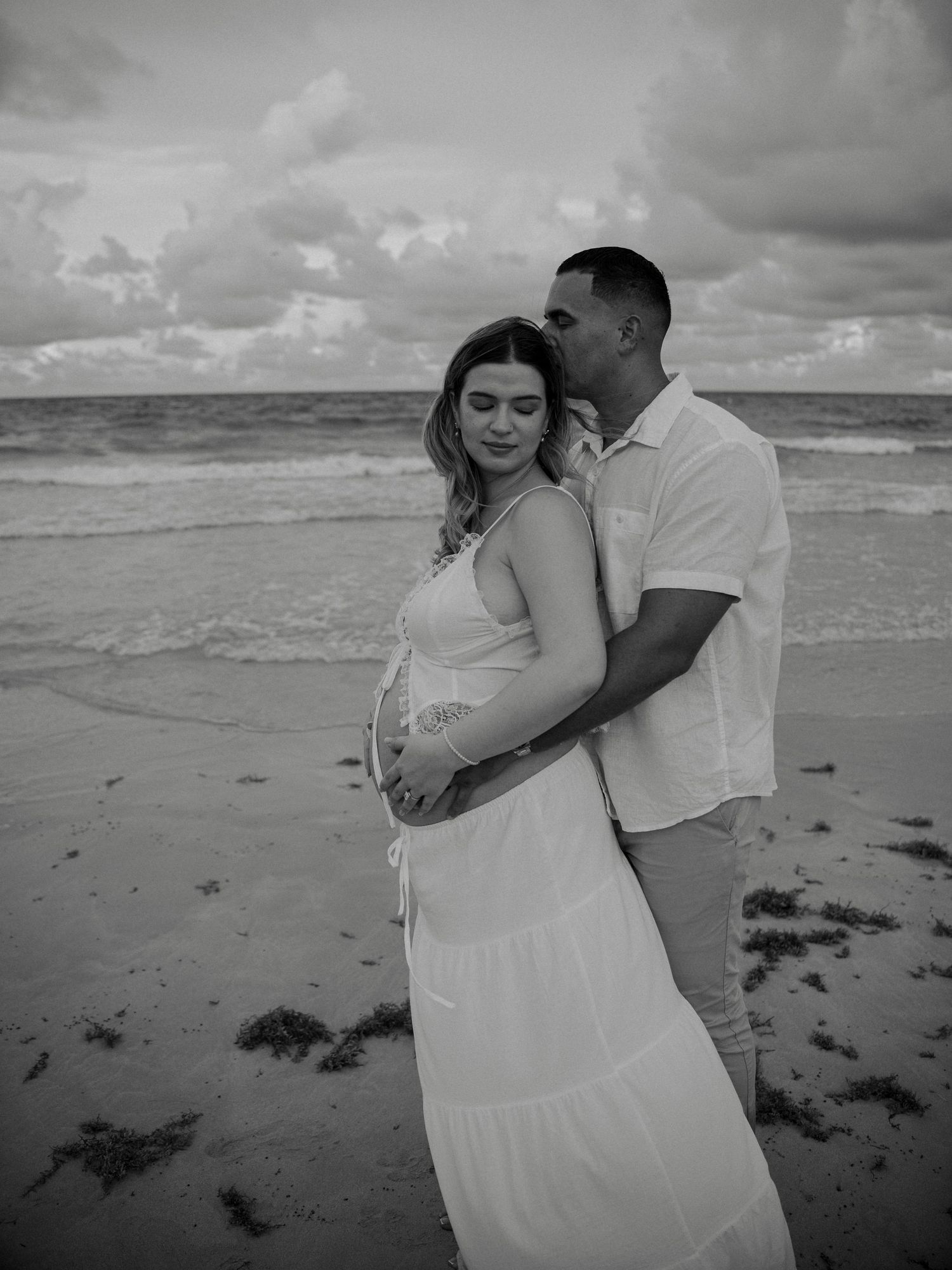 Black and white maternity photo of couple on beach, man kissing woman's head while embracing her baby bump.