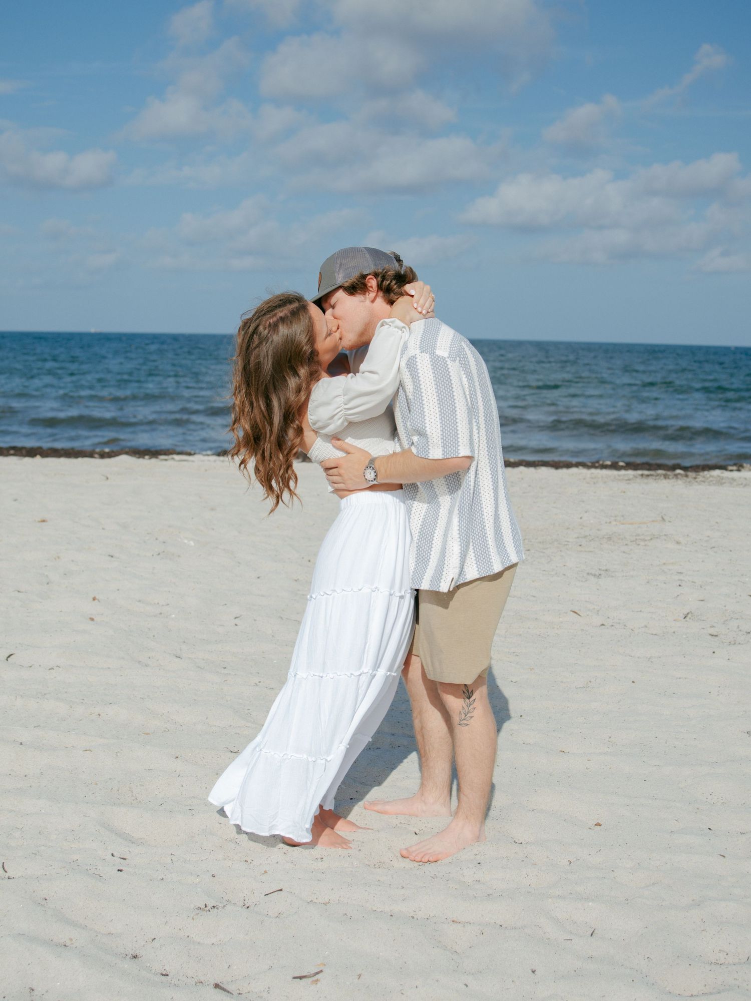Romantic couple kissing on white sand beach, woman in white maxi skirt and man in striped shirt.