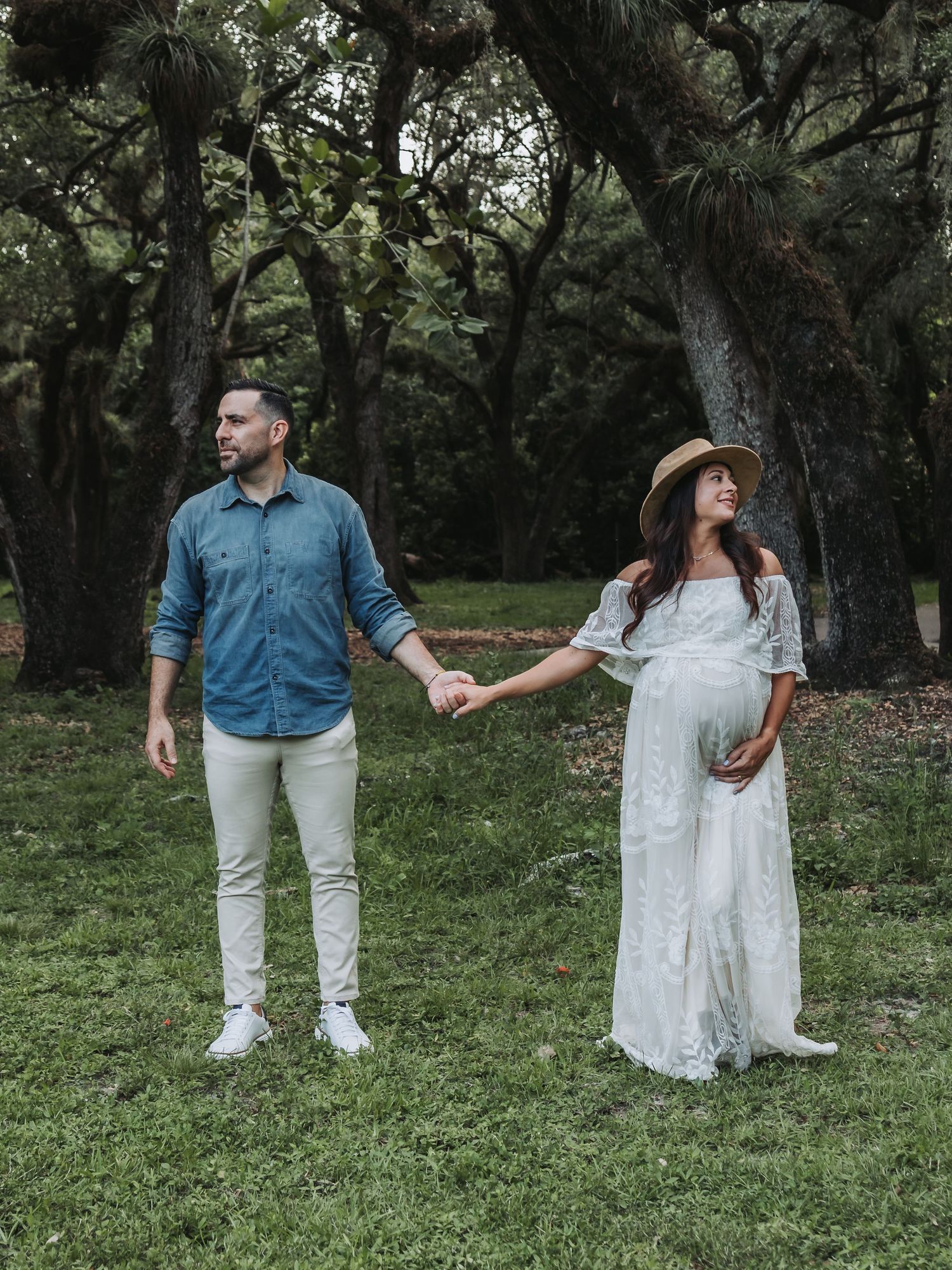 A pregnant woman in a white lace dress holds hands with her partner among towering oak trees.