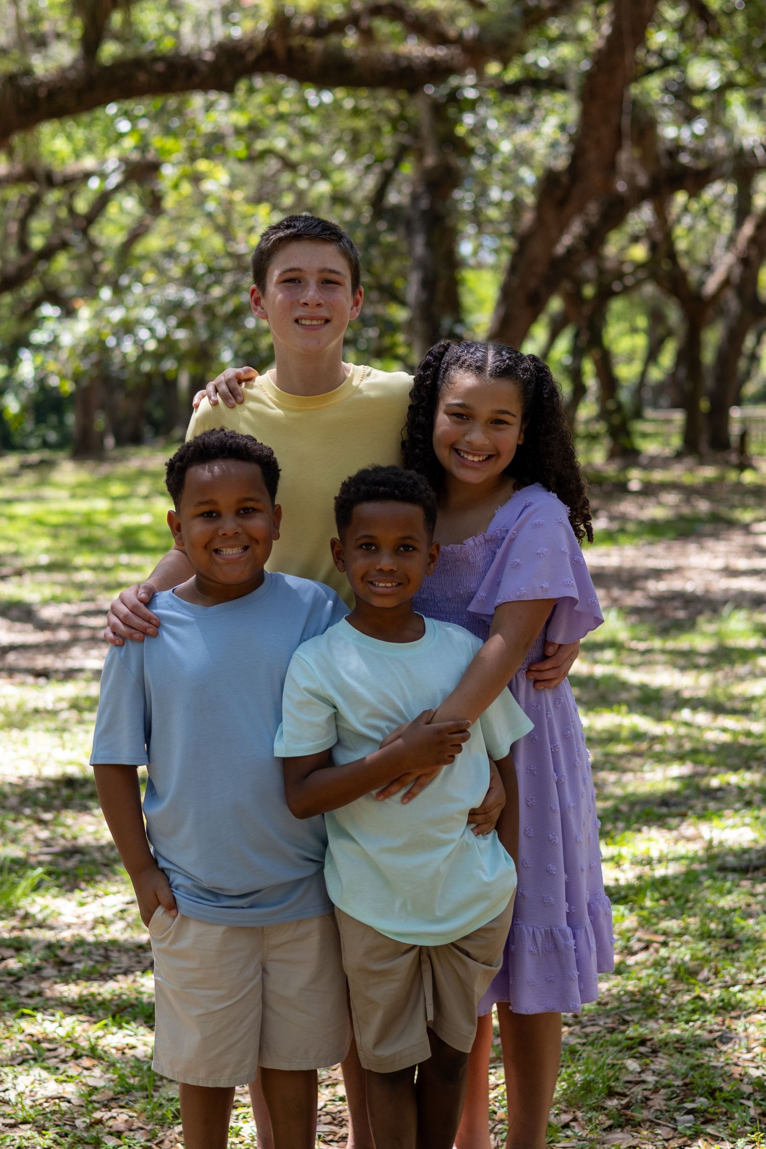 Four siblings embracing and smiling together in a lush green park on a sunny afternoon.